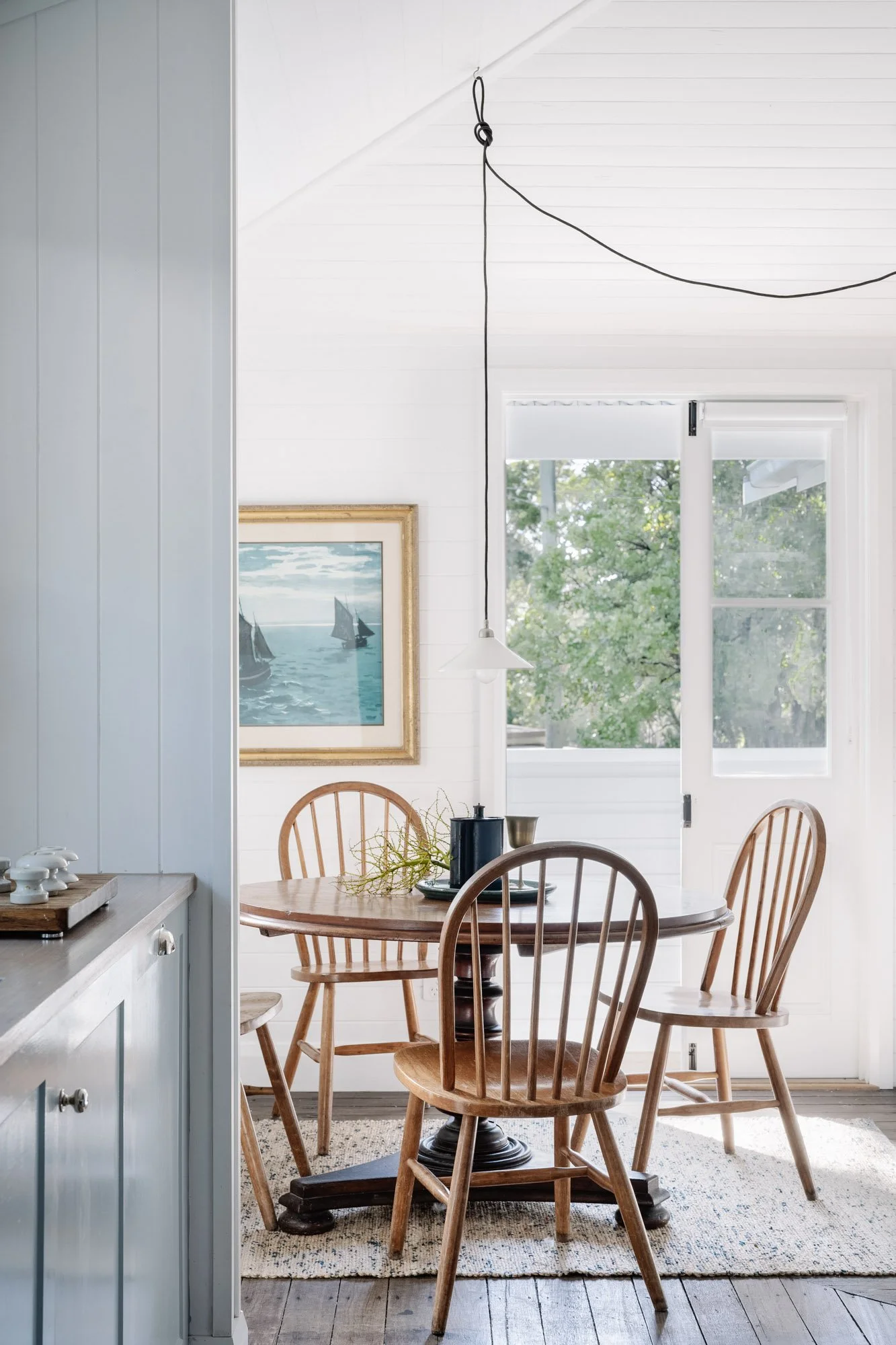 A dining area with a wooden table surrounded by four wooden chairs, set on a light patterned rug, with a white pendant light hanging above. A picture of sailboats on the water hangs on the white shiplap wall, and a large window and glass door let in natural light and views of greenery outside.