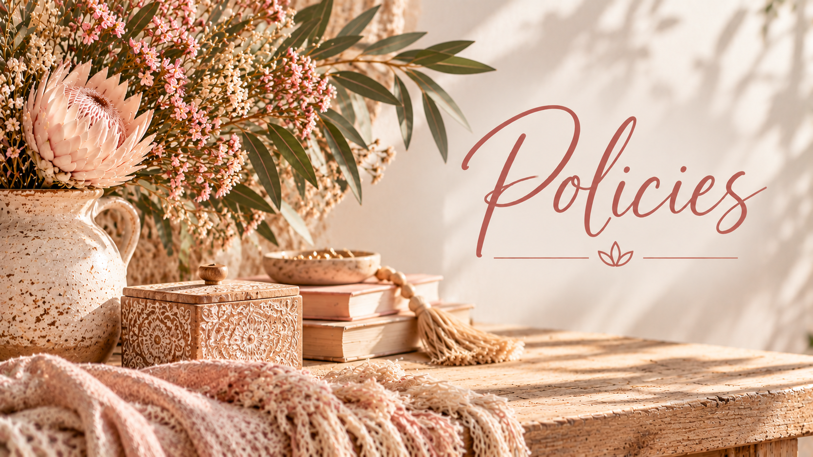A rustic wooden table decorated with a large cream-colored ceramic vase filled with pink protea and other pink flowers and greenery, a patterned wooden jewelry box, a small bowl, and a stack of books with a tassel bookmark. The background features shadowed leaves and the word 'Policies' written in elegant script.