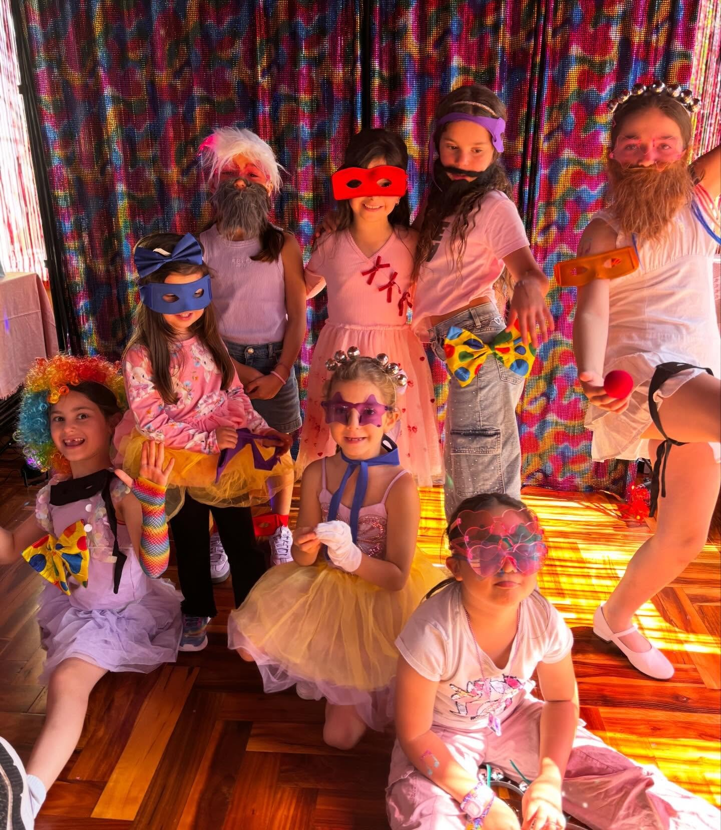 Children dressed in colorful costumes and masks, smiling and posing indoors against a multicolored backdrop, possibly at a costume party or Halloween event.