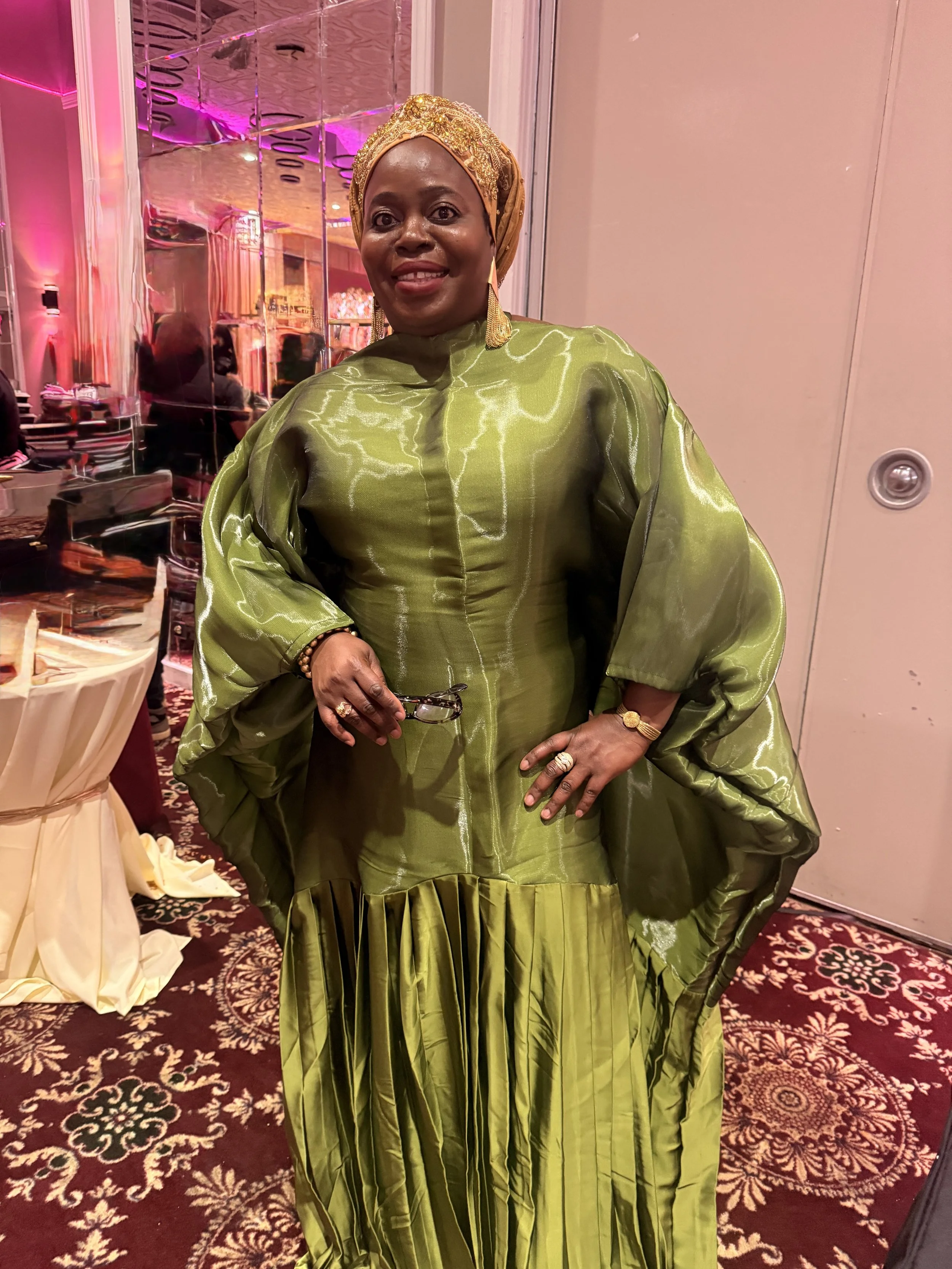 A woman dressed in green traditional African attire with gold jewelry and headwrap, standing in a decorated event hall with pink and purple lighting.