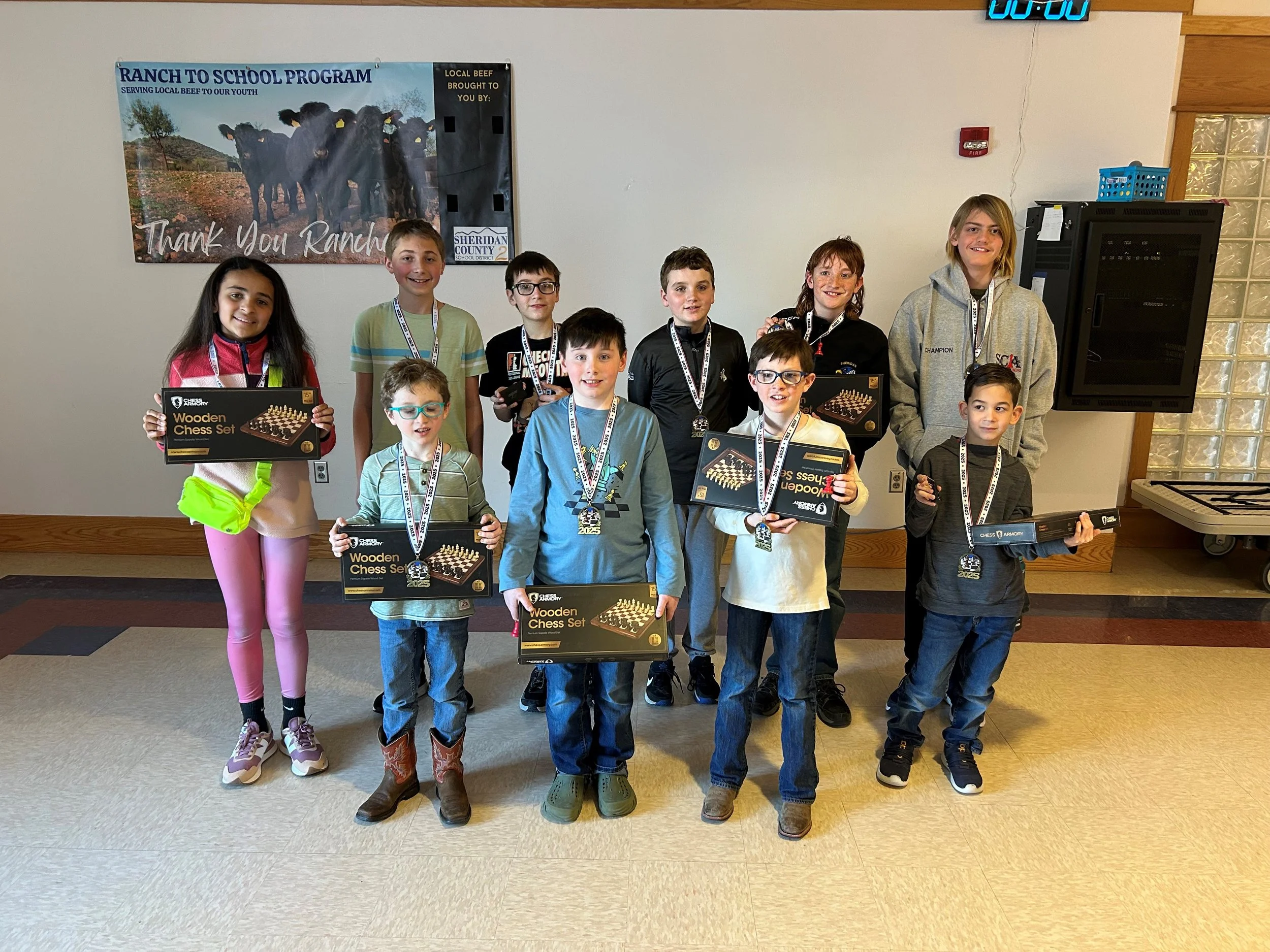 A group of children standing indoors, smiling, and holding chess sets and trophies. Behind them is a poster thanking ranchers for local beef, and there is a clock on the wall.