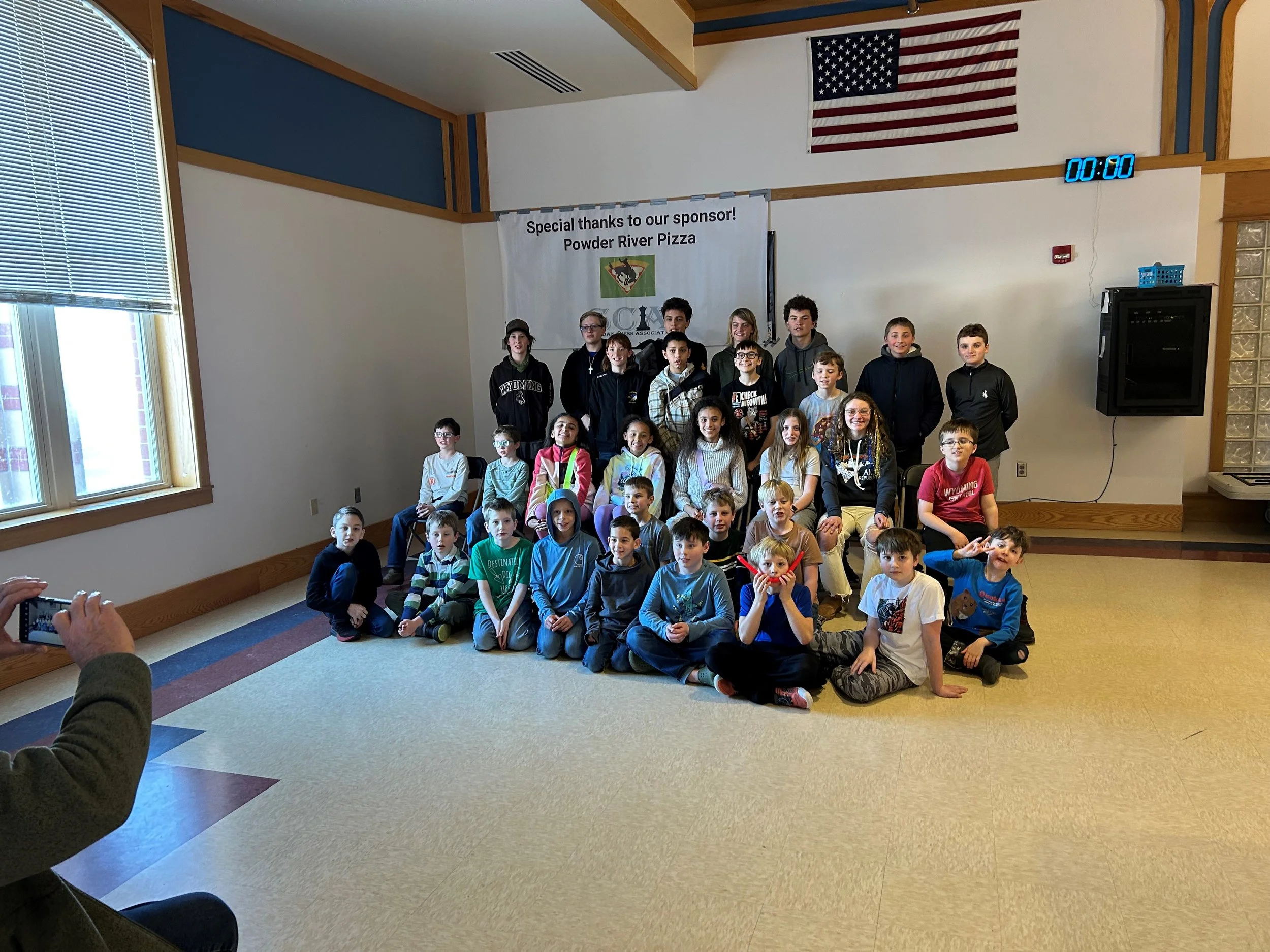 Group of children and teenagers posing for a photo inside a room with a banner that reads 'Special thanks to our sponsor! Powder River Pizza,' a clock showing 00:00, and an American flag on the wall.