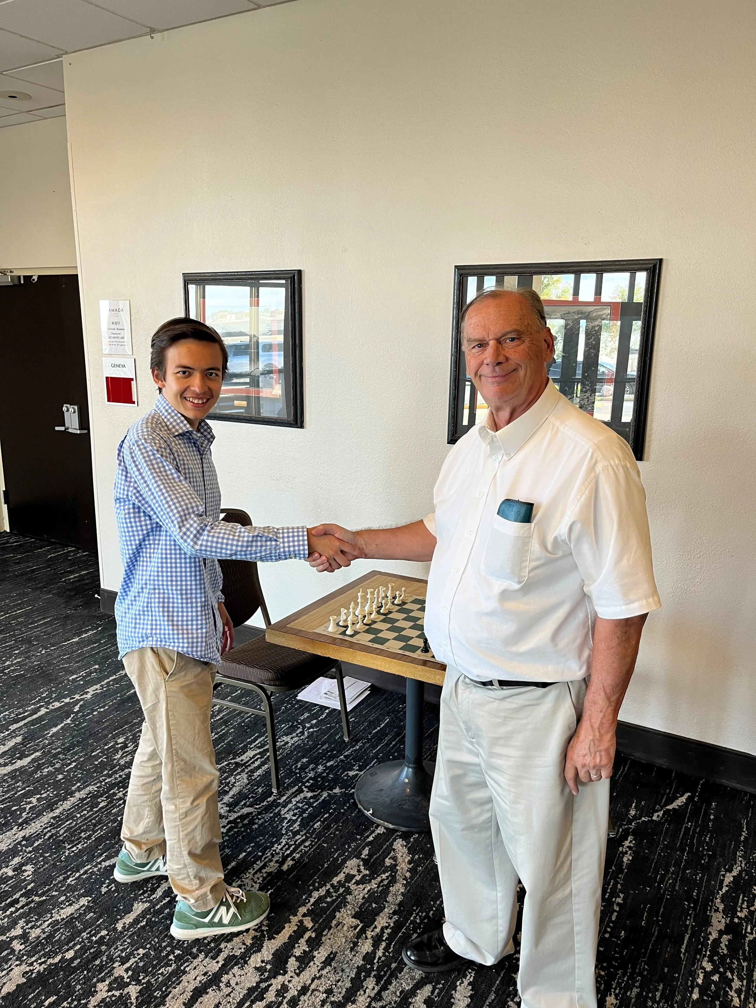A young man and an older man shaking hands in front of a chessboard at a table, smiling. The young man wears a blue checkered shirt and beige pants, the older man wears a white shirt and light-colored pants. The background features two framed windows on a beige wall and a black and white patterned carpet.