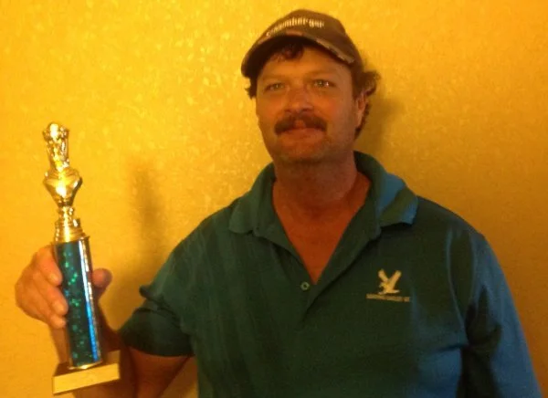 Man with a mustache and cap holding a trophy, standing against a yellow wall.