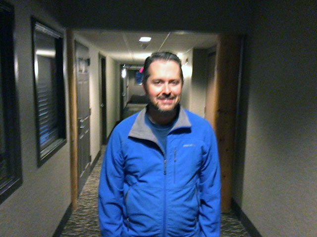 A man with long hair, beard, and mustache wearing a blue jacket standing in a corridor with doors on each side.
