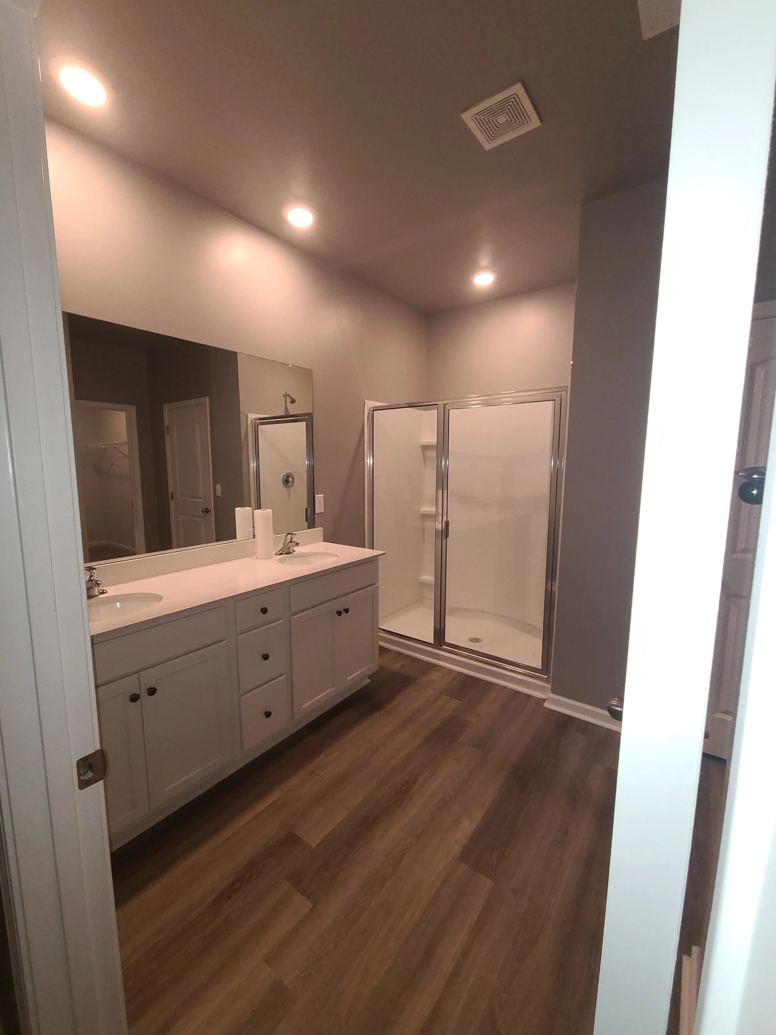 A bathroom with a double sink vanity, a mirror, a glass shower enclosure, and wood-look flooring.