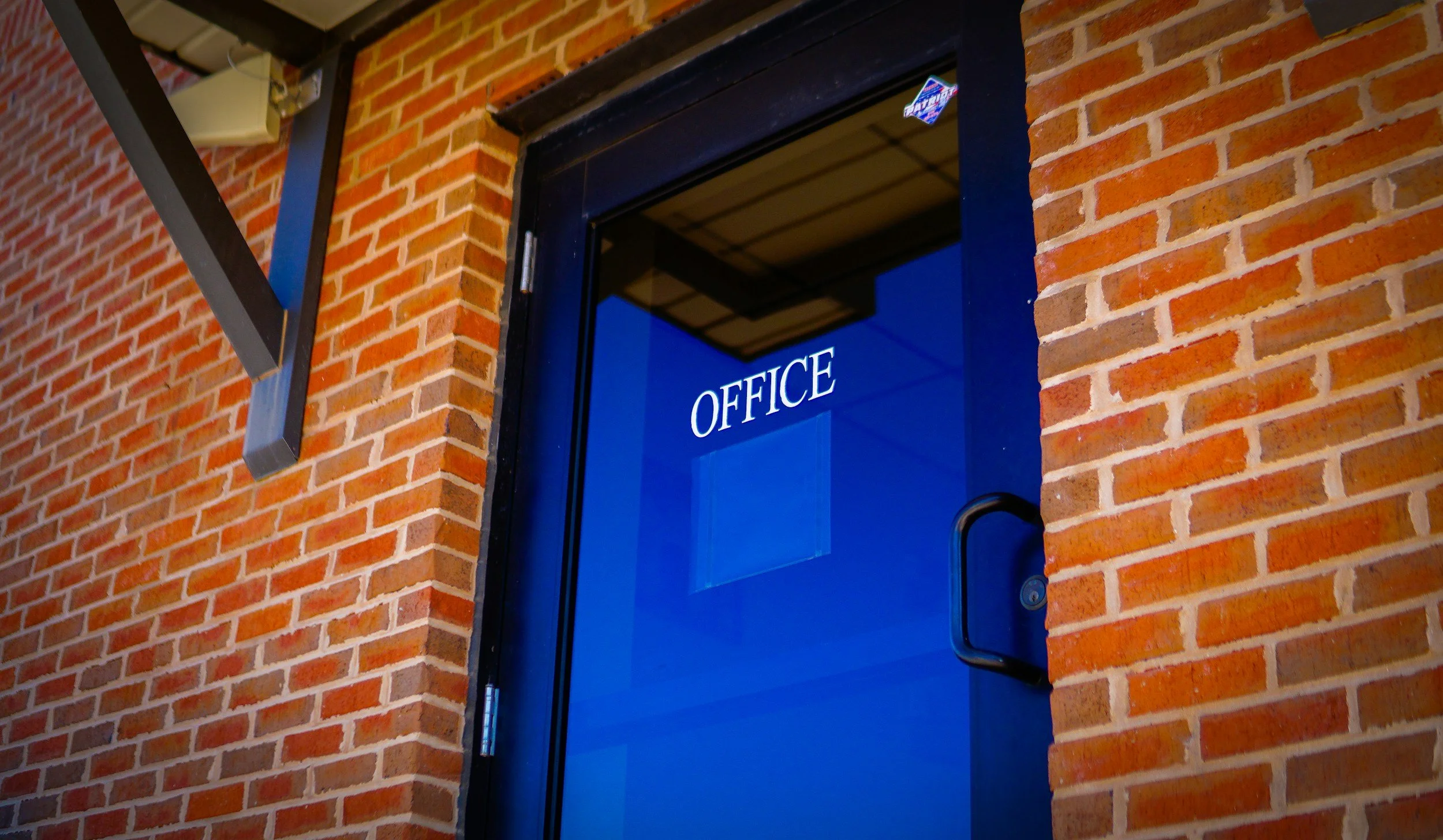Blue door with a glass window that has the word 'OFFICE' written on it, set into a red brick wall.