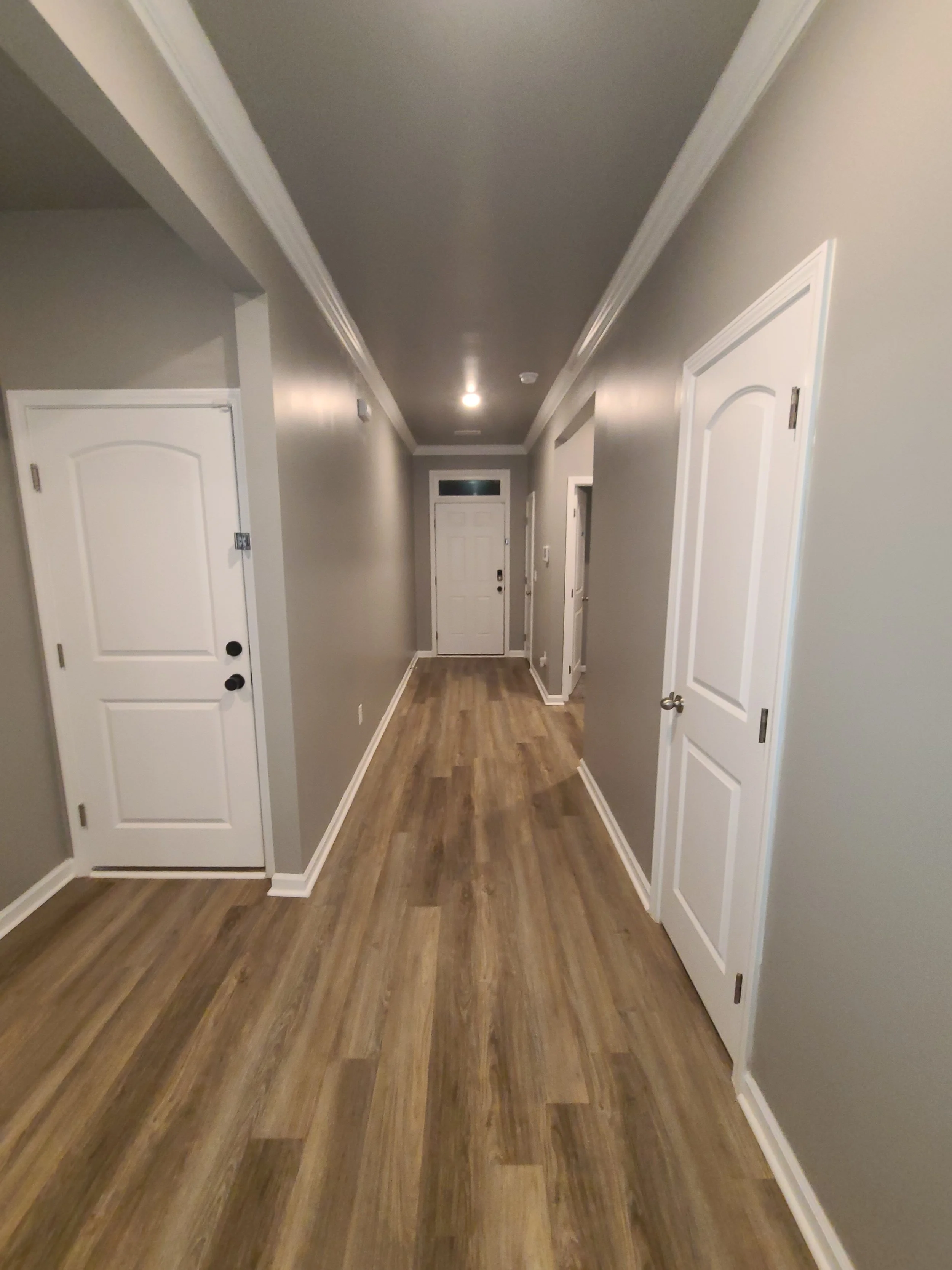 A hallway with gray walls, white doors, and wood laminate flooring, leading to a front door at the end.