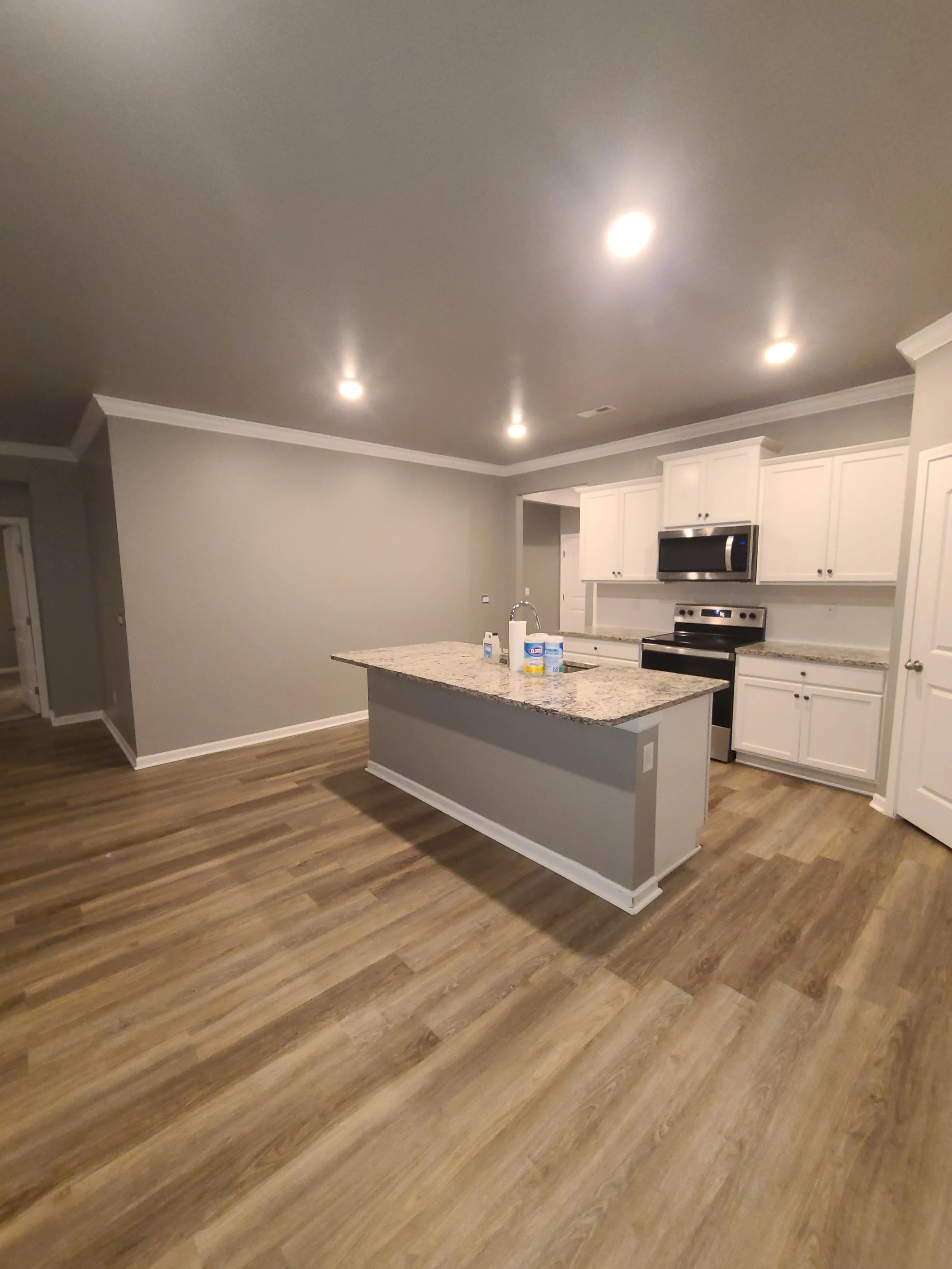 The image shows a modern kitchen with white cabinets, stainless steel appliances, a granite countertop island, and wood-look flooring. Recessed ceiling lights illuminate the space.