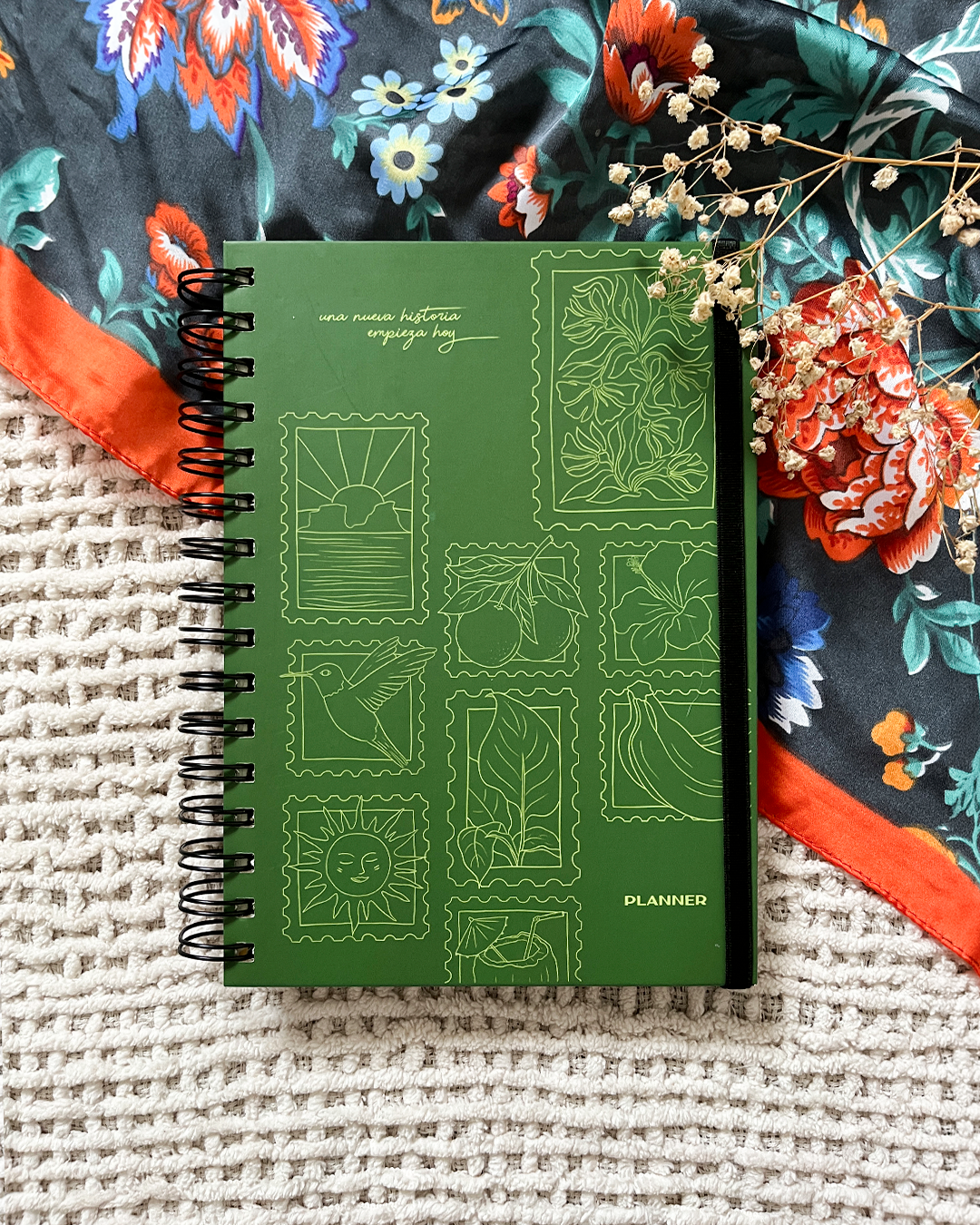 Un cuaderno de planificación de color verde con citas en español y dibujos de estampillas, rodeado de tela con estampado floral y flores secas blancas y rojas.