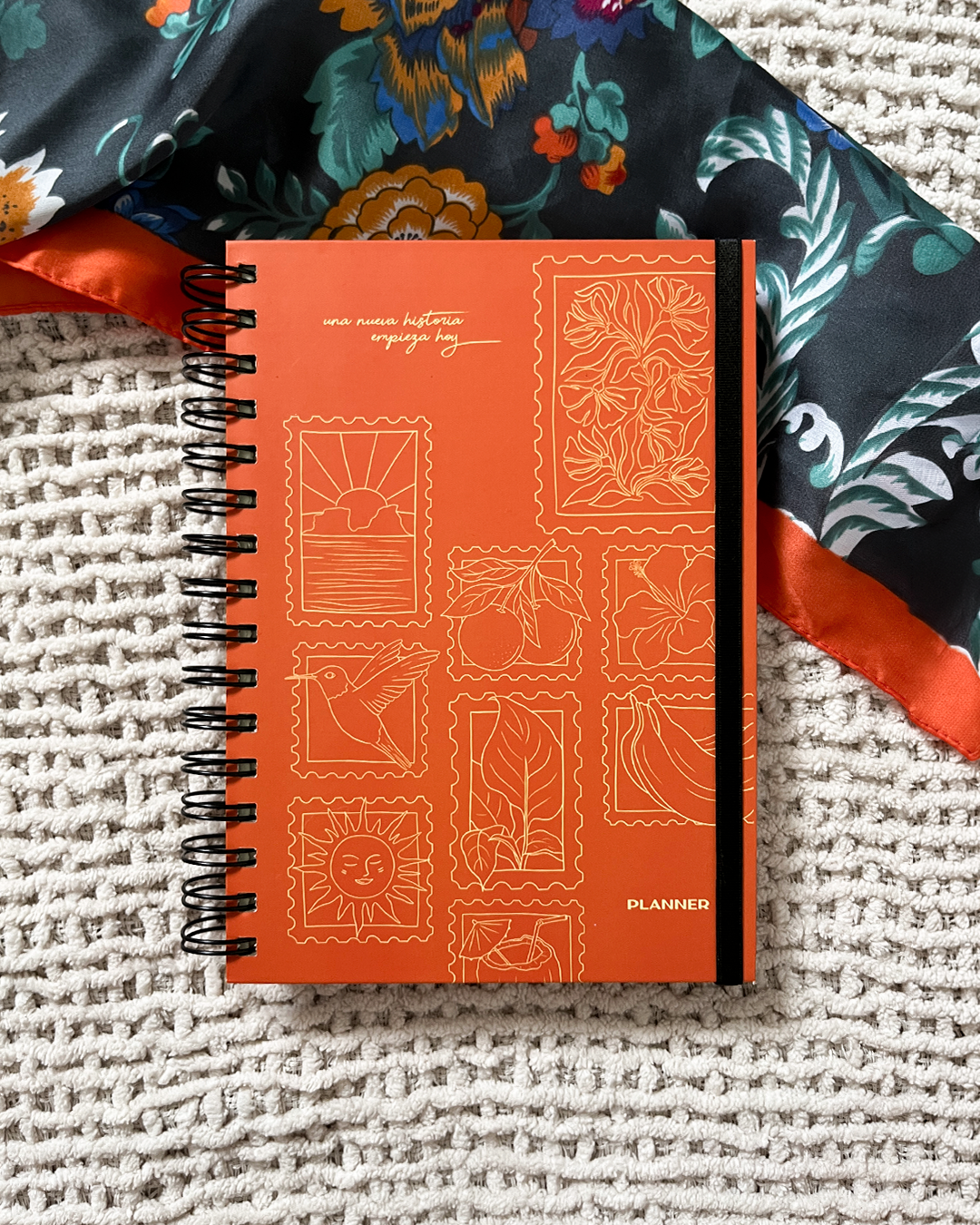 Una libreta de planificador de color naranja con ilustraciones de sellos de varias imágenes, sobre una superficie de alfombra. Parte de un paño con estampado floral y banda color naranja en la esquina superior izquierda.