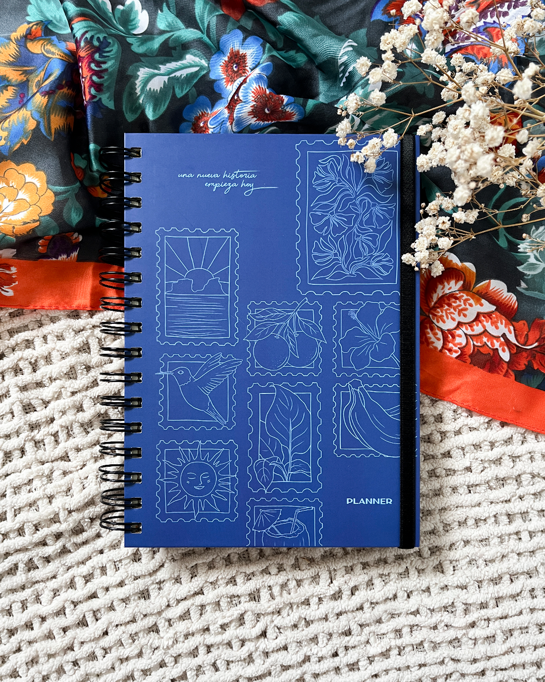 Una agenda azul con ilustraciones de sellos, rodeada de telas coloridas con estampados florales y unas flores blancas secas.