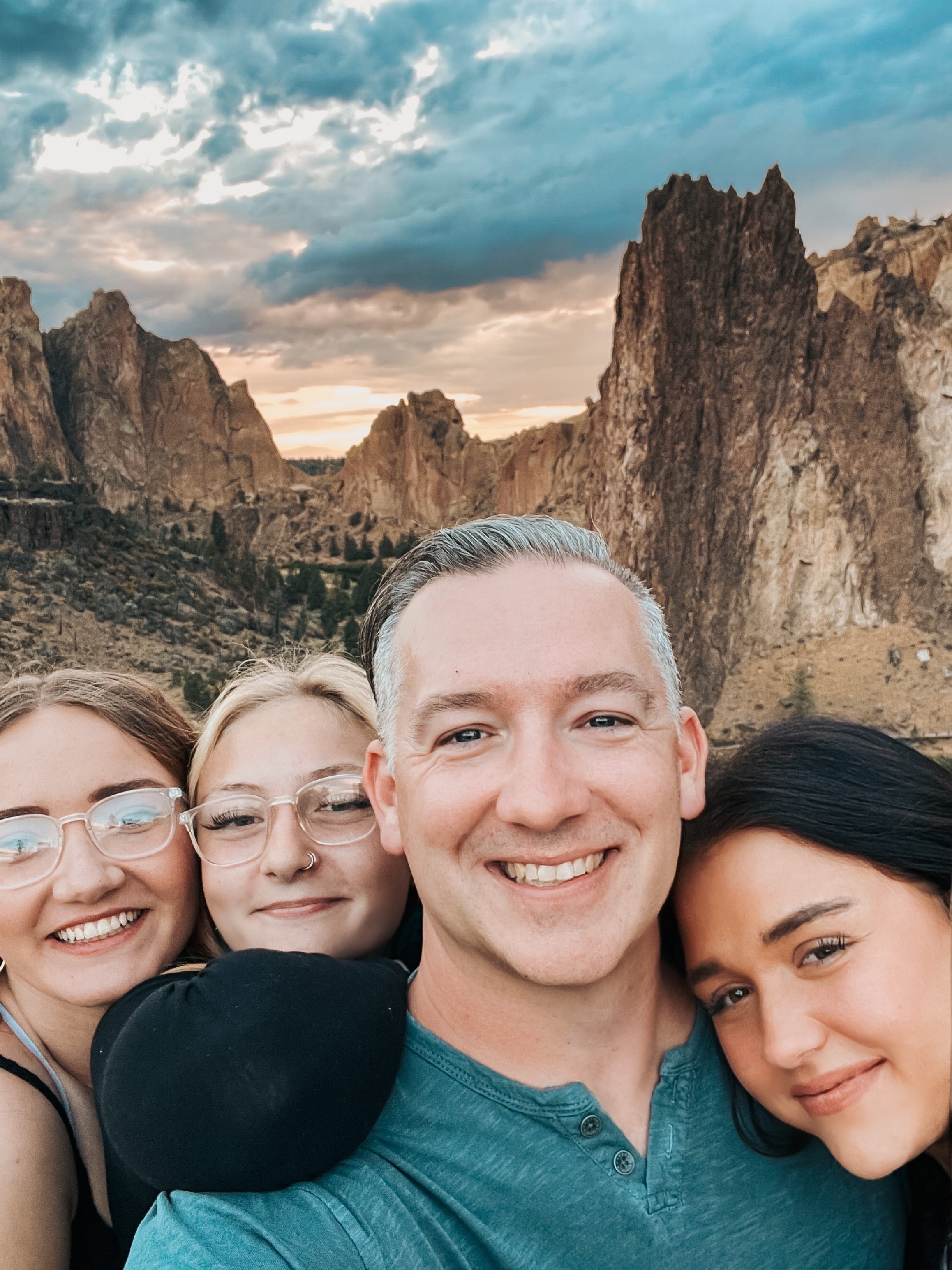 Smiling group of people outdoors, with rocky mountains and cloudy sky in the background.