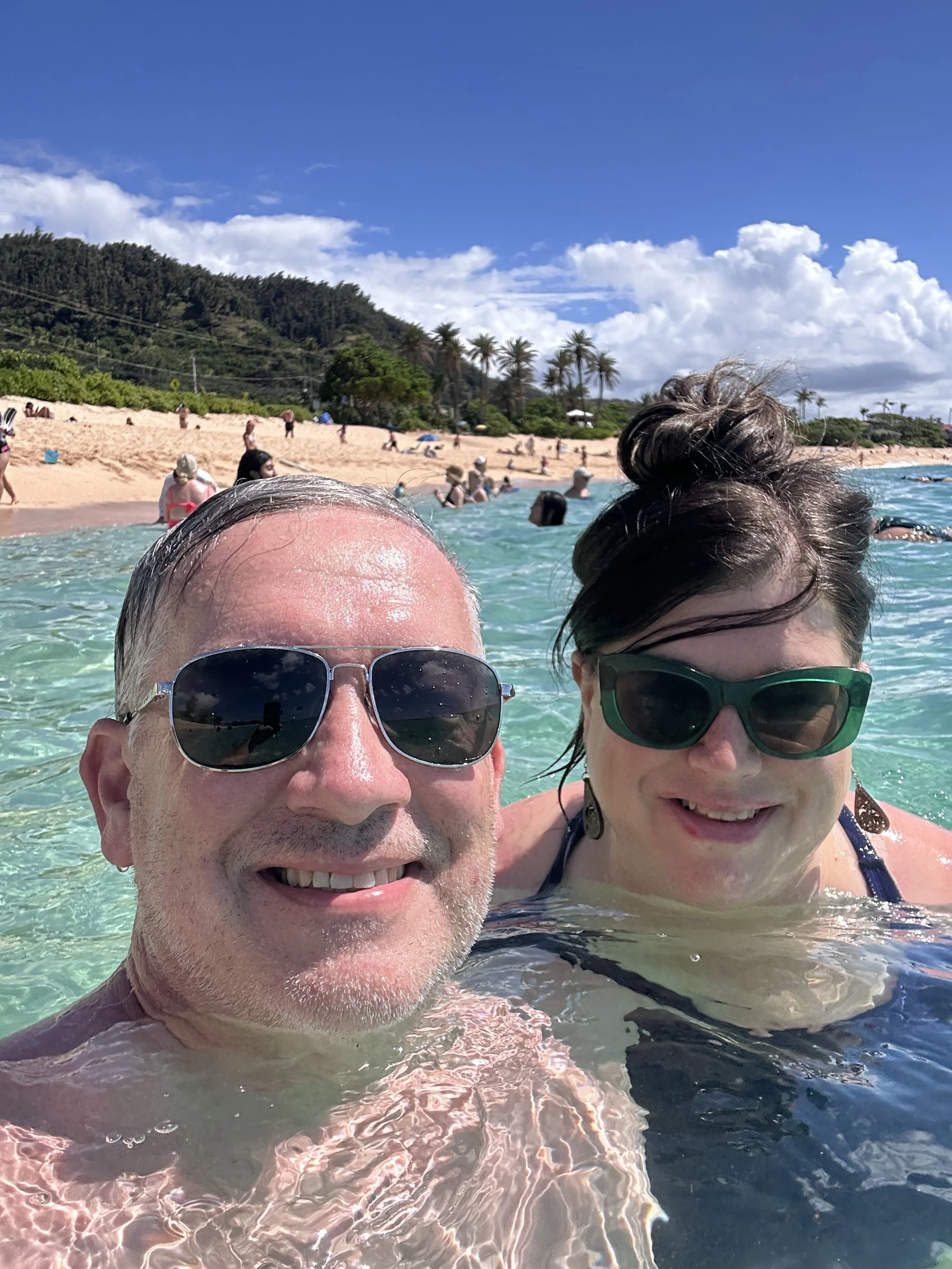 Two people with sunglasses enjoying a swim at the beach, with a sandy shore, palm trees, and a partly cloudy sky in the background.