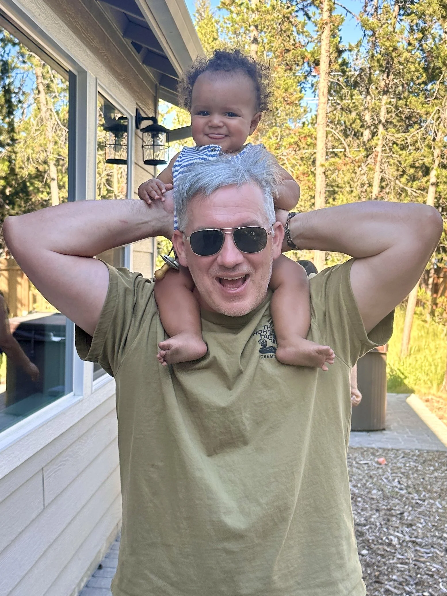 A man with gray hair, wearing sunglasses and a tan t-shirt, carries a smiling baby on his shoulders outside a house with trees in the background.