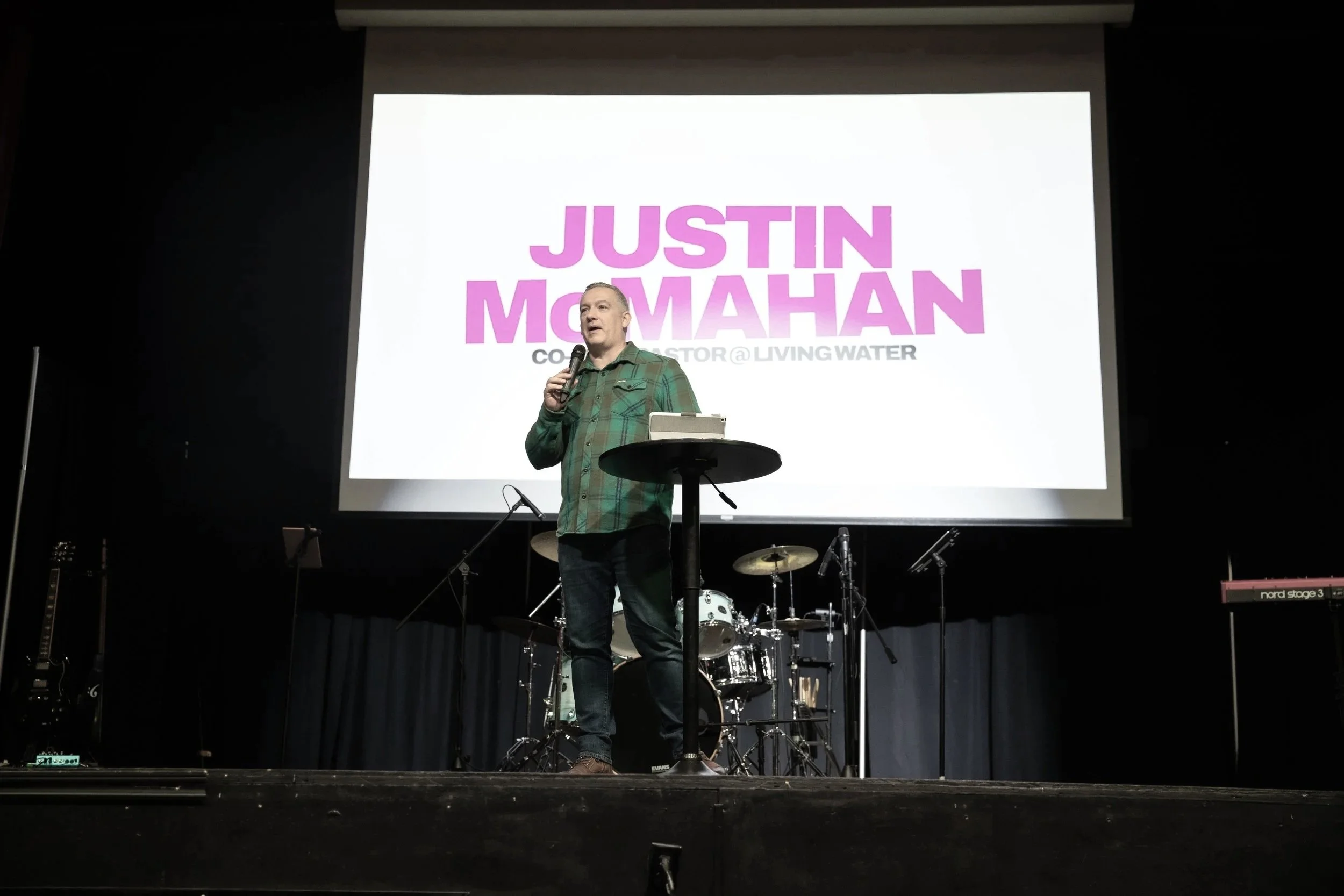 A man in a green plaid shirt speaking into a microphone on a stage with musical instruments, large screen behind him displaying the name Justin Moomahan and the title 'Co-Founder @ Living Water'.