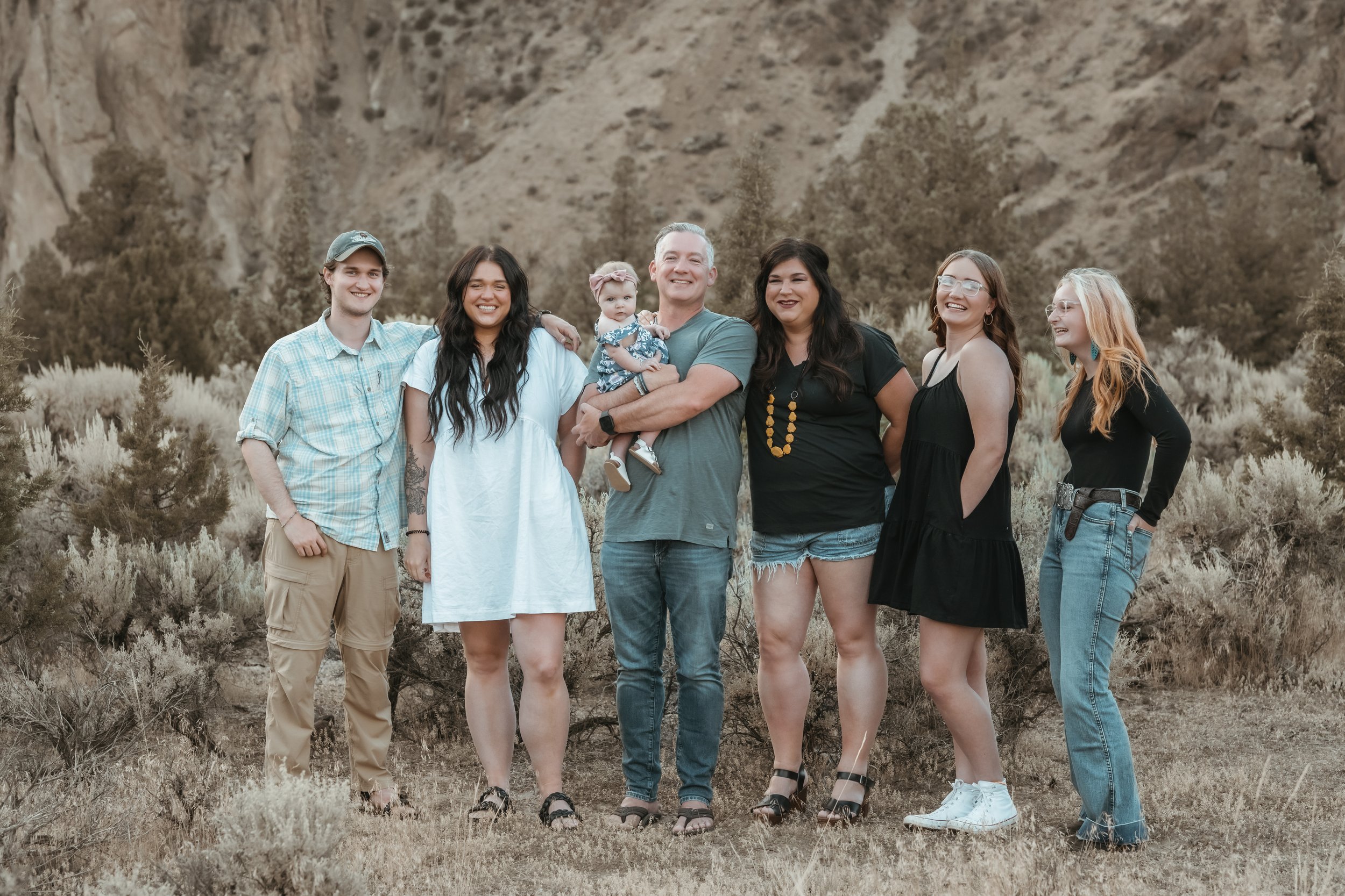 A group of seven people, including a baby, standing outdoors in front of a rocky hillside. The group is smiling and posing together, with some standing close and others with arms around each other. They are casually dressed, with variations in clothi