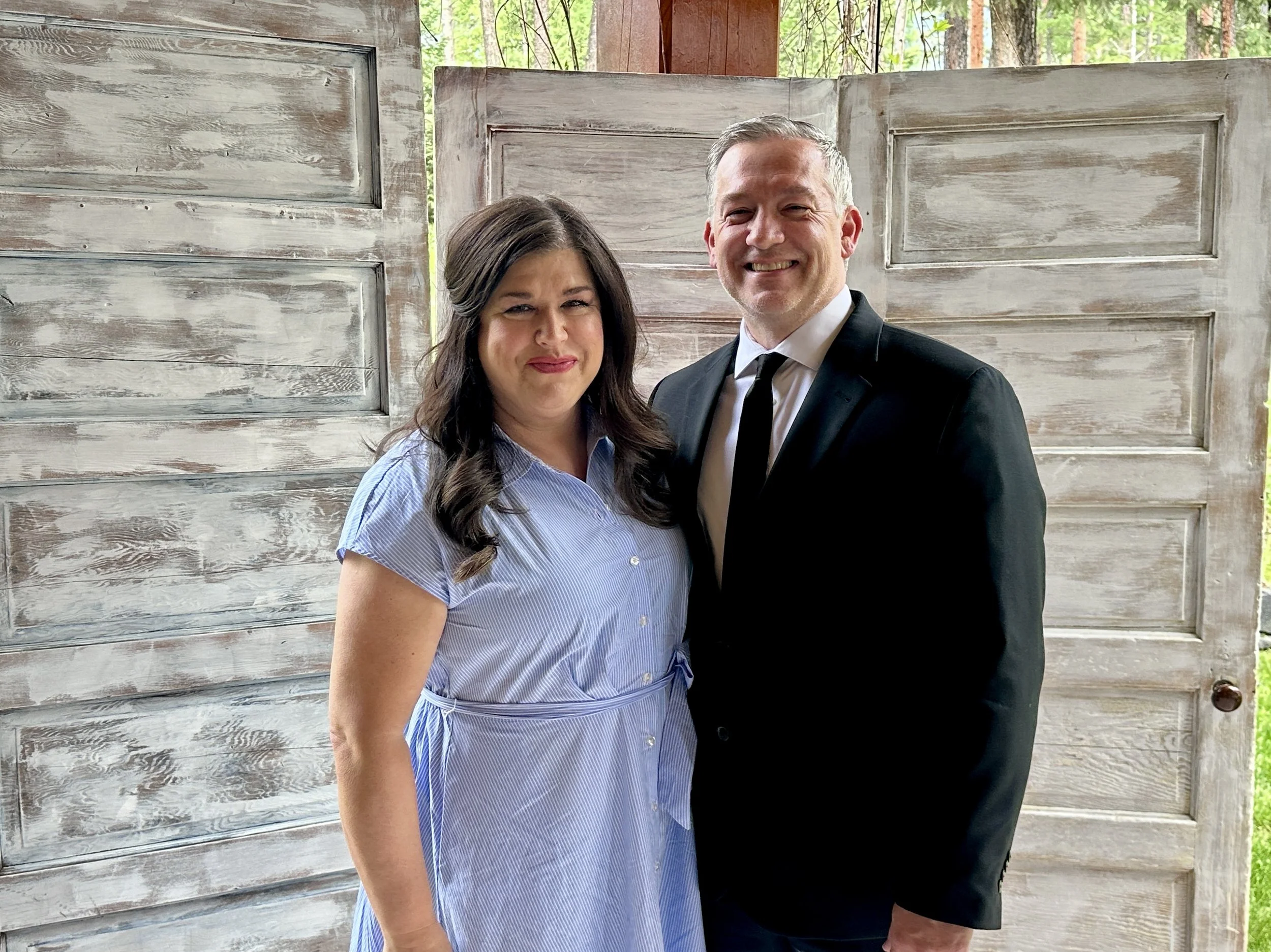 A man and woman smiling in front of a rustic wooden backdrop, with trees visible in the background.