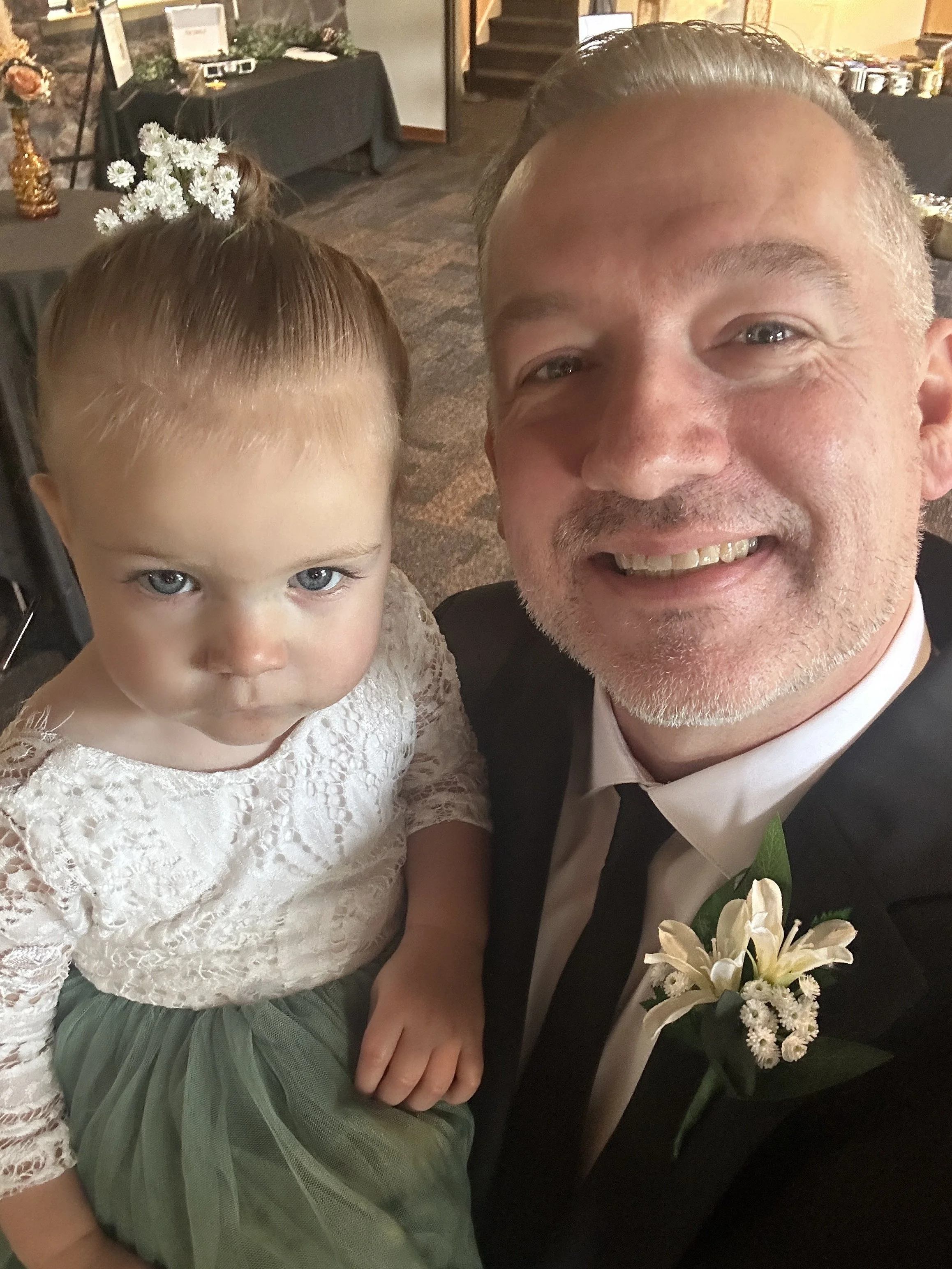 A smiling man in a black suit with a white shirt and black tie takes a selfie with a young girl. The girl has light blue eyes, light skin, and brown hair styled in a top bun with white flowers. She wears a white, lace dress with a green tulle skirt. 