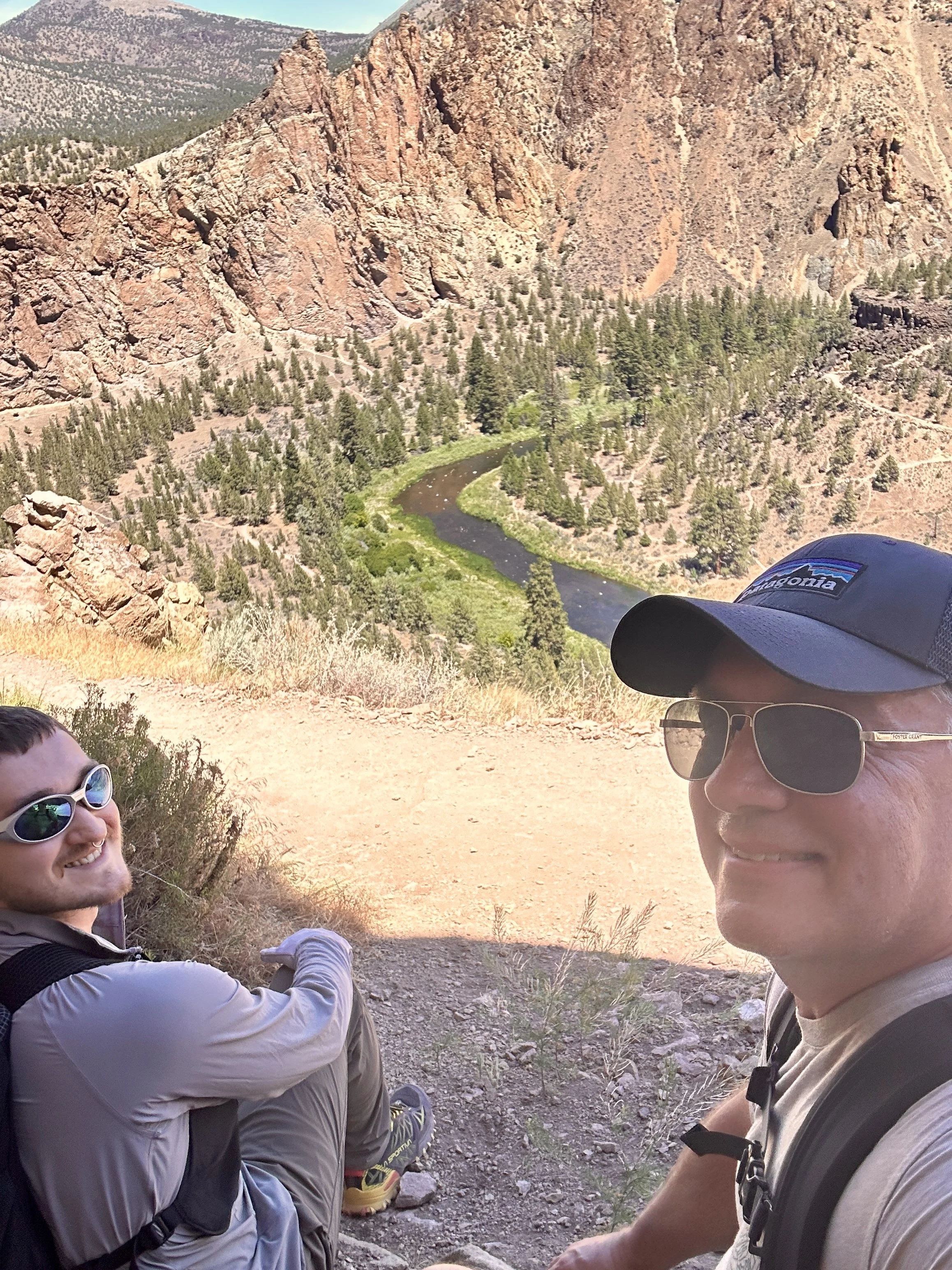 Two people hiking in a canyon with rocky cliffs, green trees, a river, and a clear sky in the background.