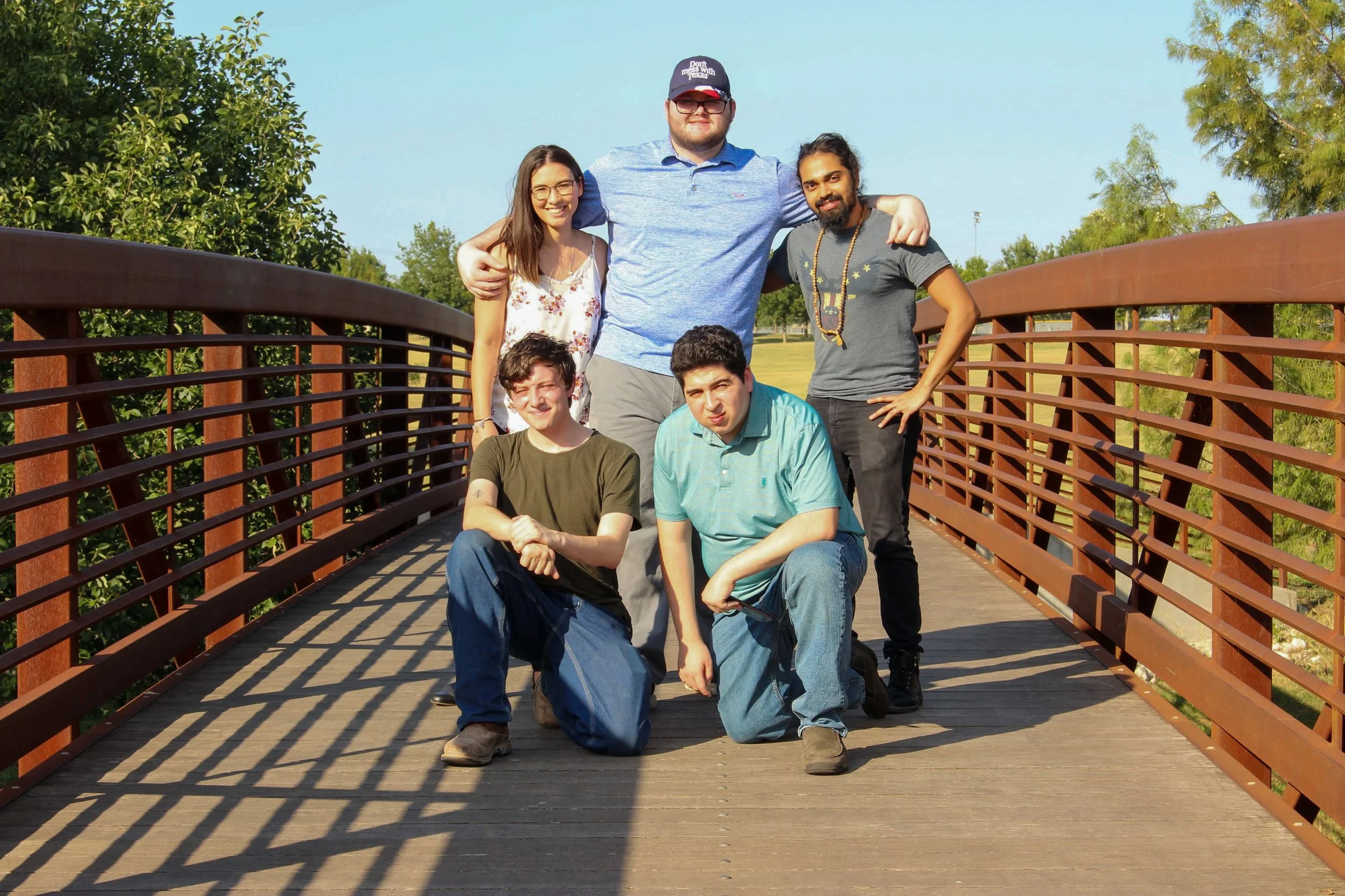Group of five young adults posing on a wooden bridge outdoors during daytime. Two are kneeling in front, three standing behind, with trees and a clear blue sky in the background.