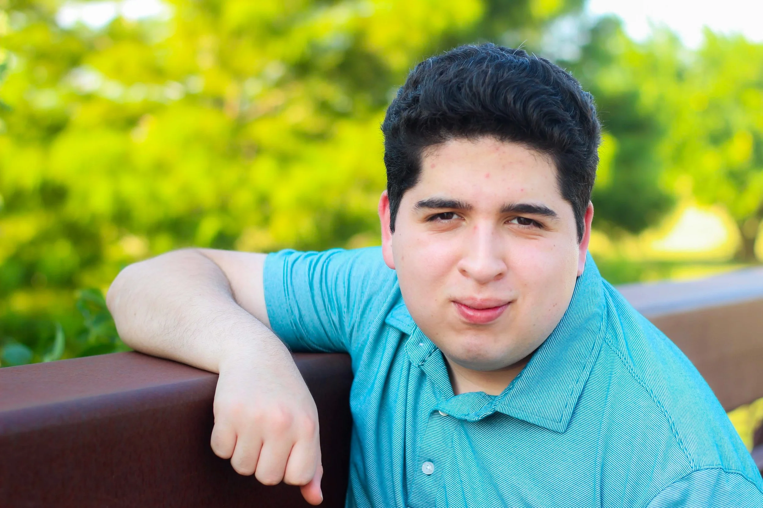 A teenage boy with dark hair and a light blue shirt resting his arm on a wooden park bench with a blurred green foliage background.