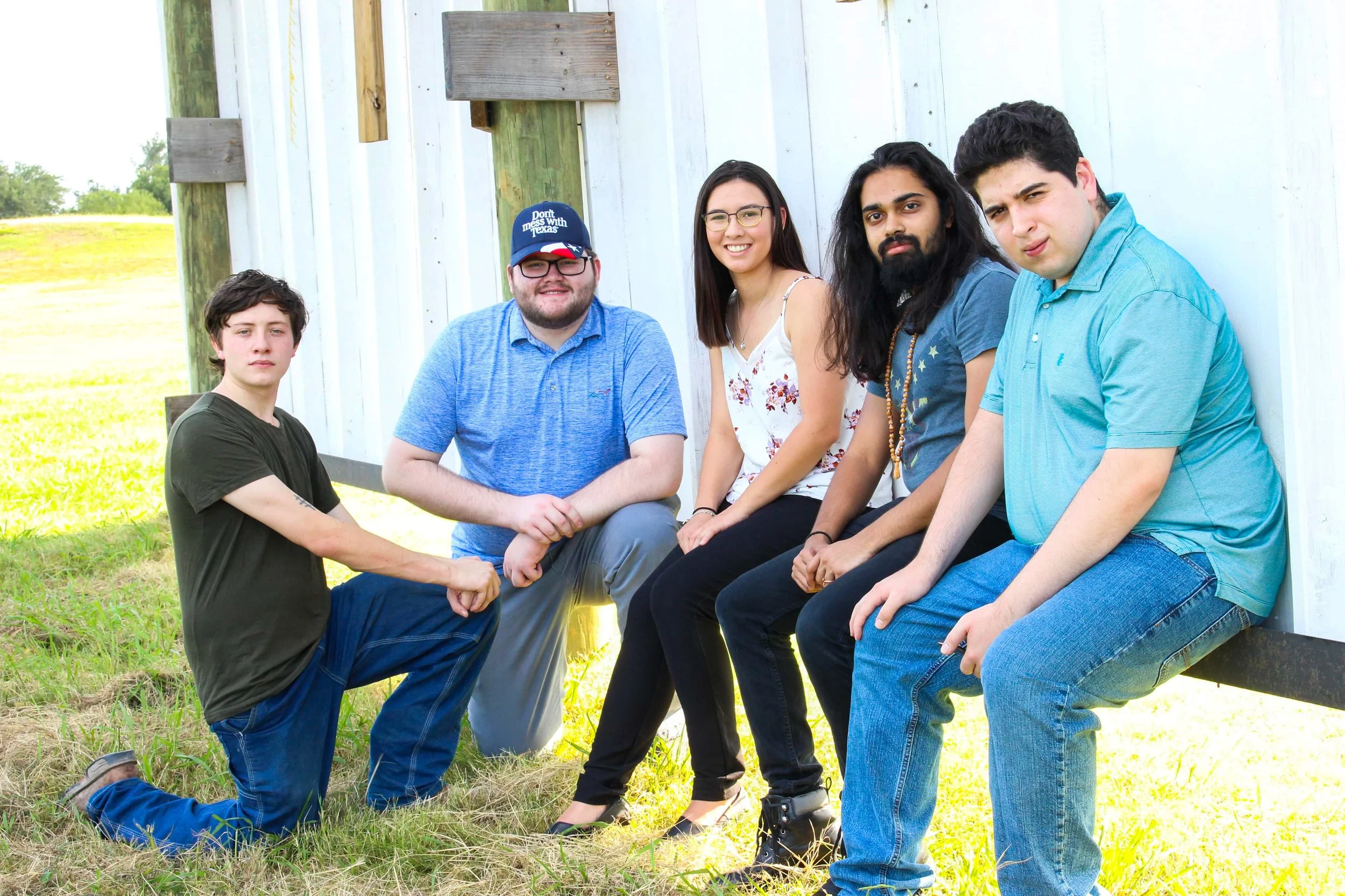 A diverse group of six people sitting and leaning against a white wooden wall outdoors on grass, smiling and looking at the camera, with a background of green trees and open sky.