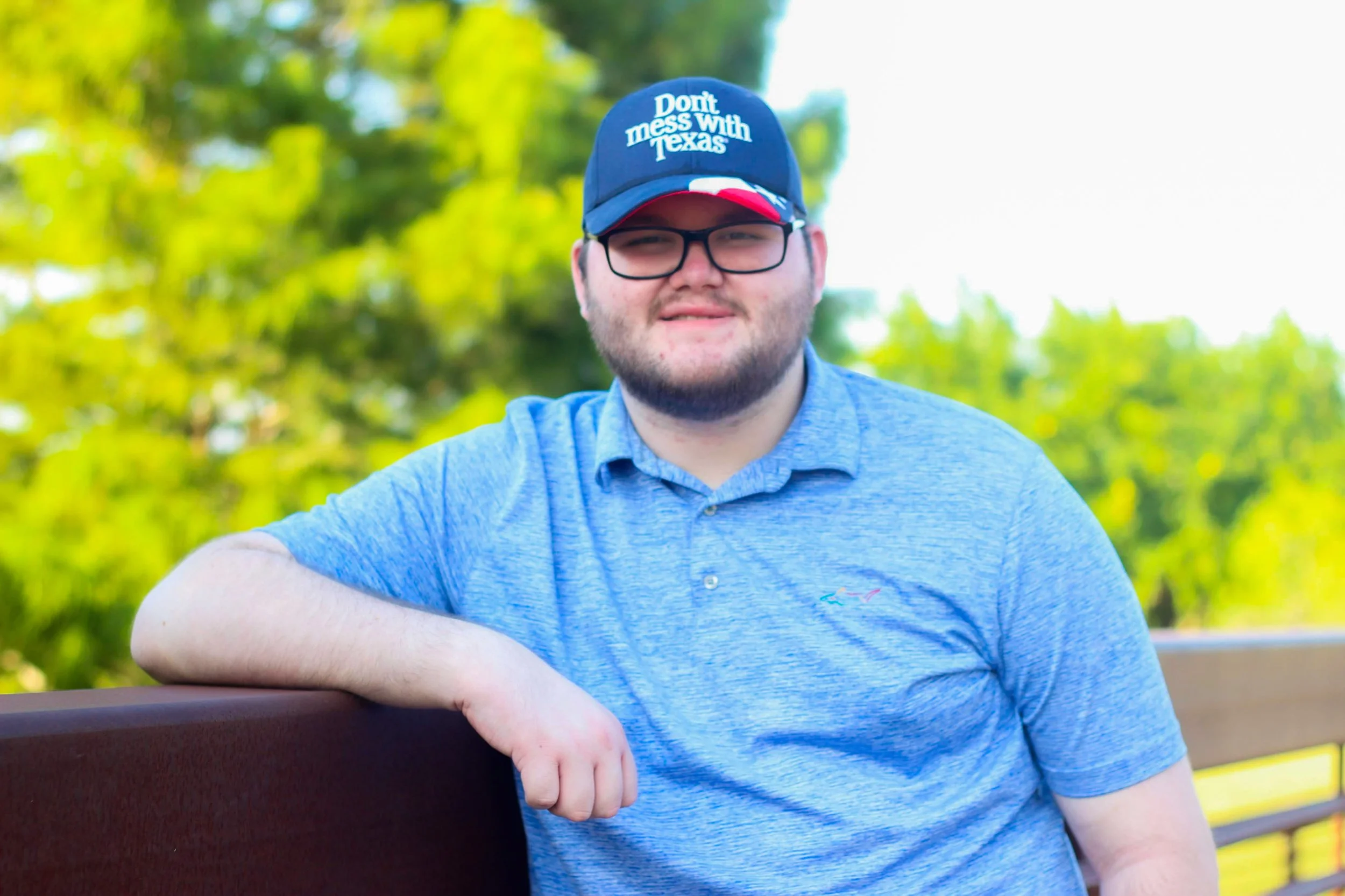 A man with glasses and a beard sitting on a park bench, wearing a blue polo shirt and a cap that says, 'Don't mess with Texas.' He is outdoors with green trees in the background.
