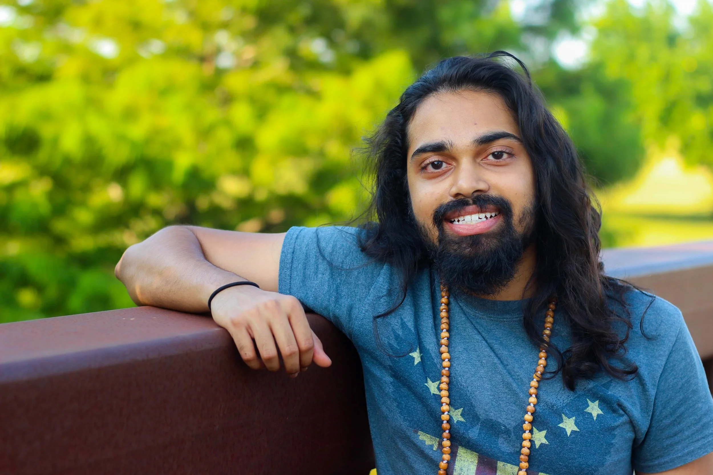 A young man with long dark hair and a beard sitting on a park bench, smiling, with a green, blurred background of trees and sunlight.
