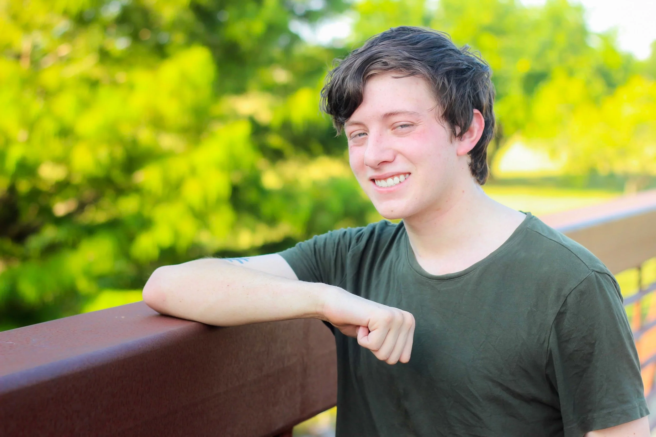 A young man with dark wavy hair sitting outdoors on a park bench, smiling slightly with his eyes partially closed, wearing a dark green T-shirt, with blurred green foliage in the background.