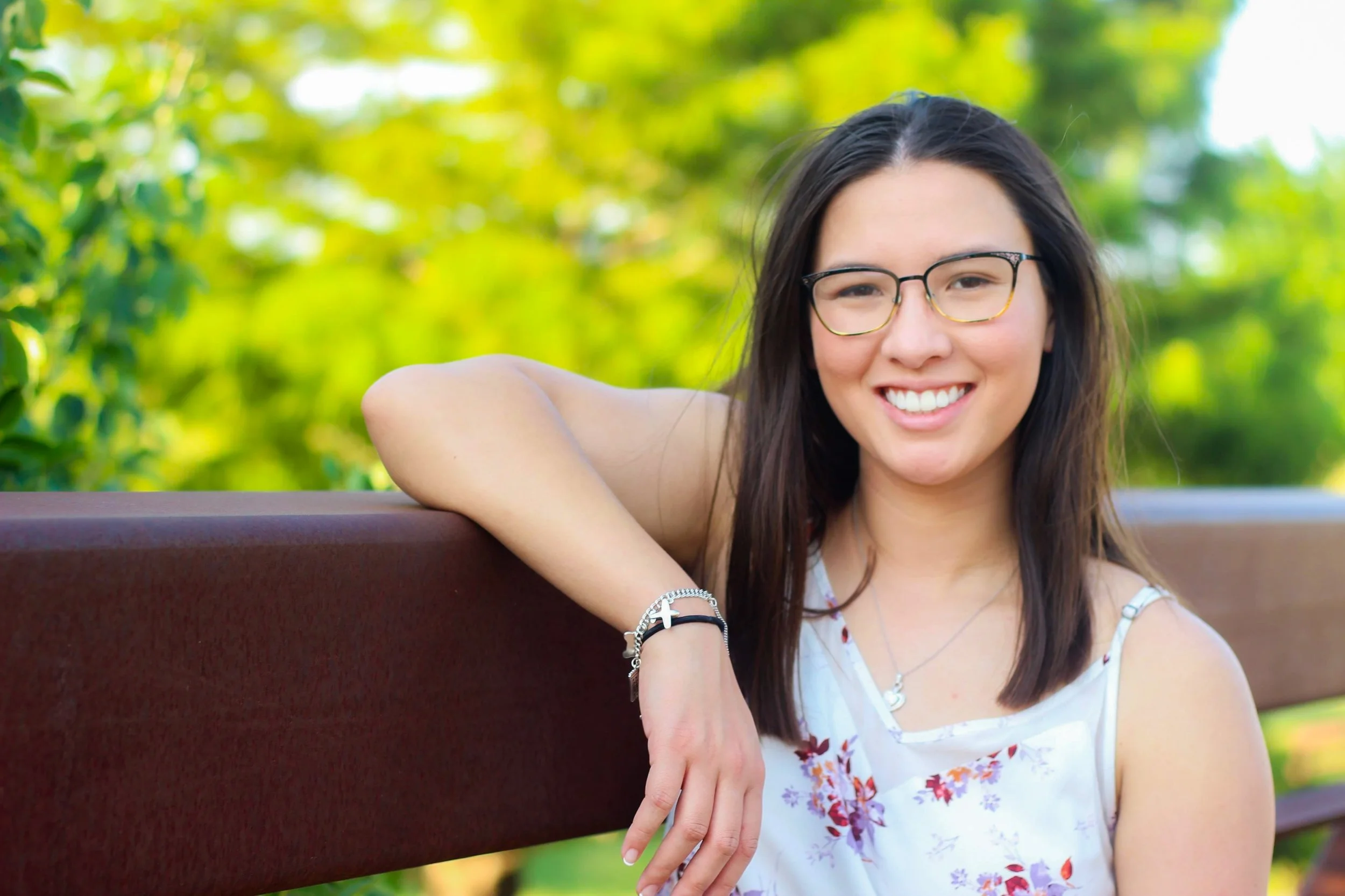 Young woman with long dark hair and glasses smiling, sitting outdoors on a bench with a green blurred background.