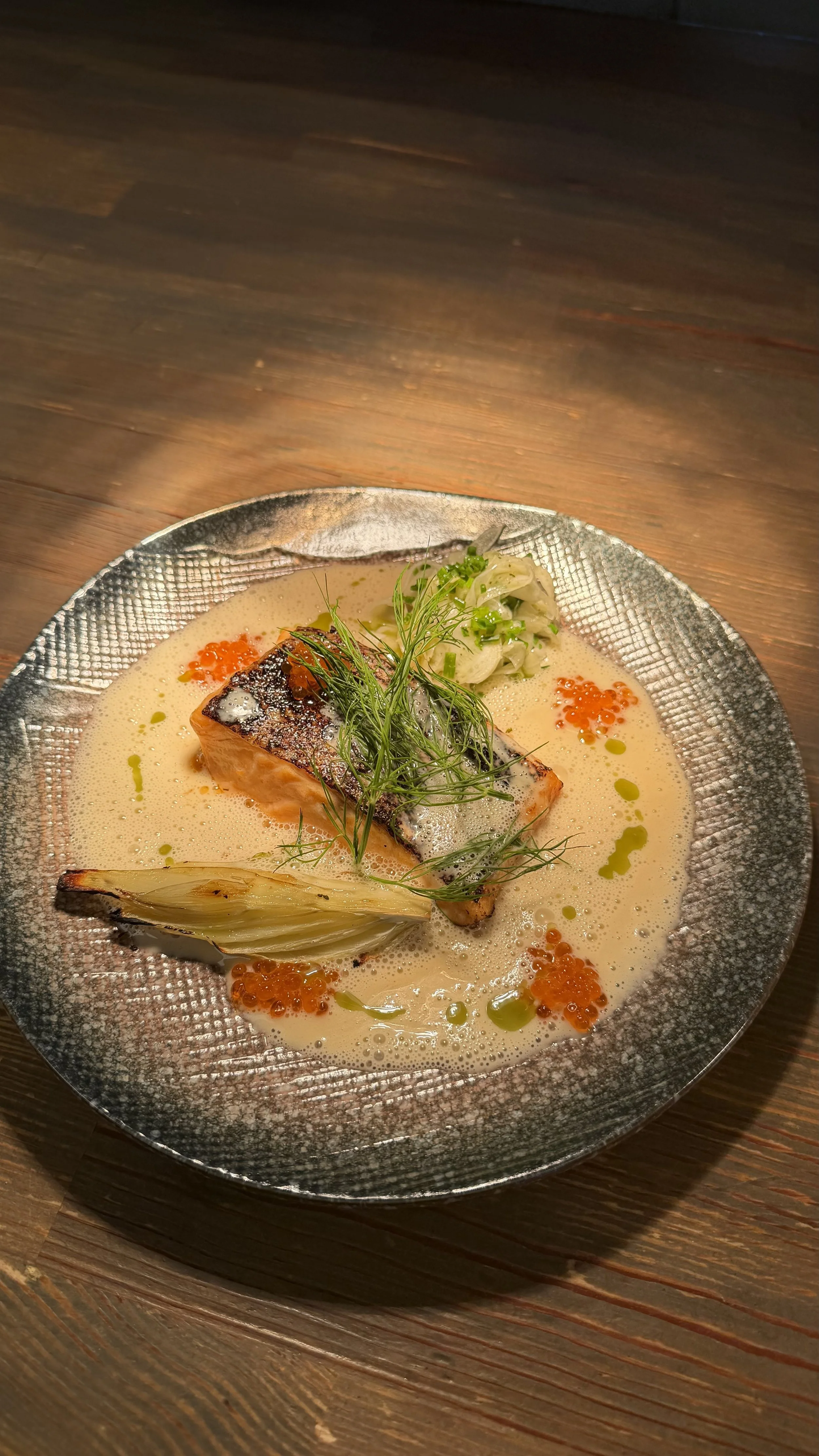 A plated gourmet dish featuring a piece of seared fish, garnished with herbs, served in a creamy sauce with orange caviar, green oil droplets, and a side of grilled fennel, on a textured silver plate.