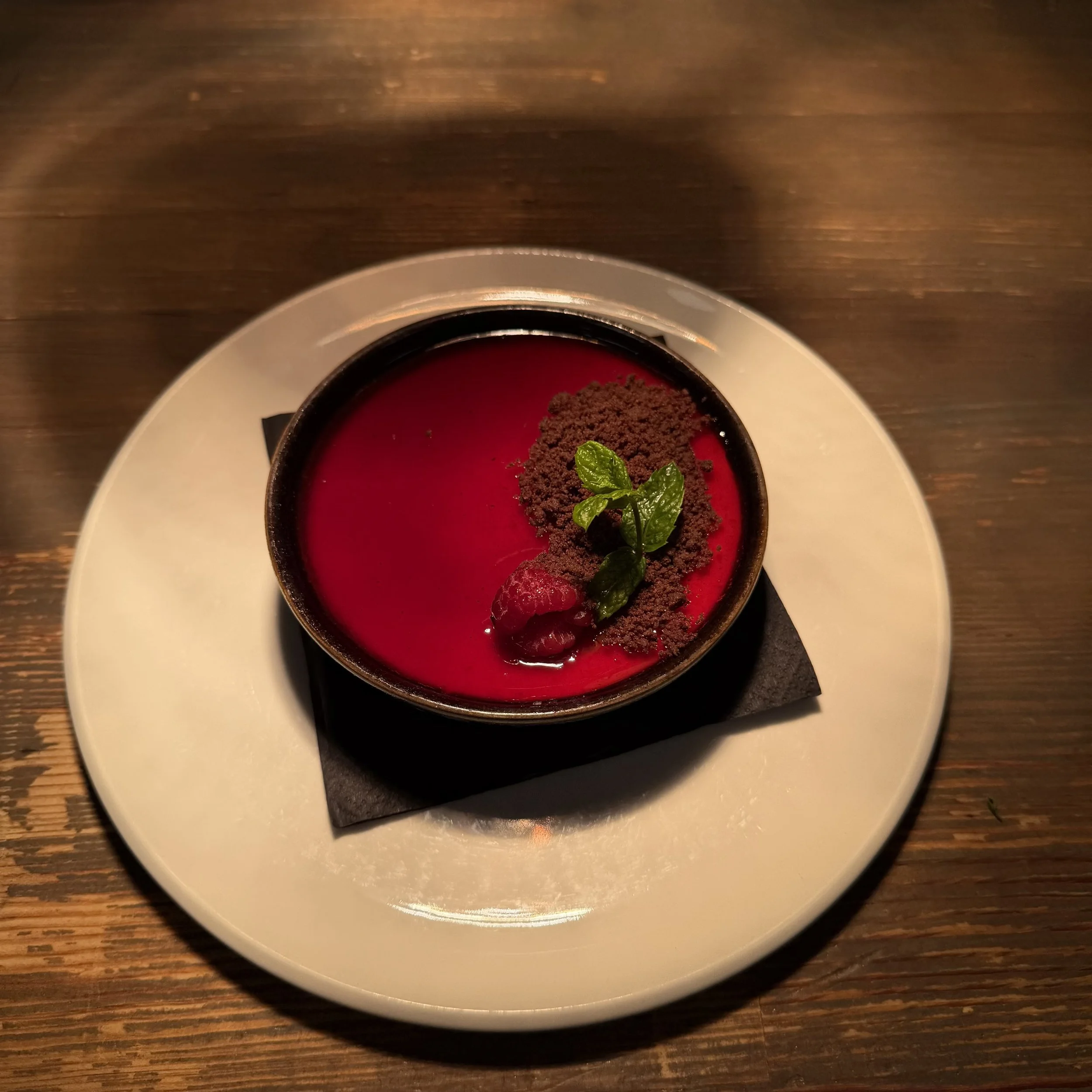 A white bowl on a white plate containing a dessert with red sauce, topped with crumbled chocolate, a raspberry, and fresh mint leaves, on a wooden table.