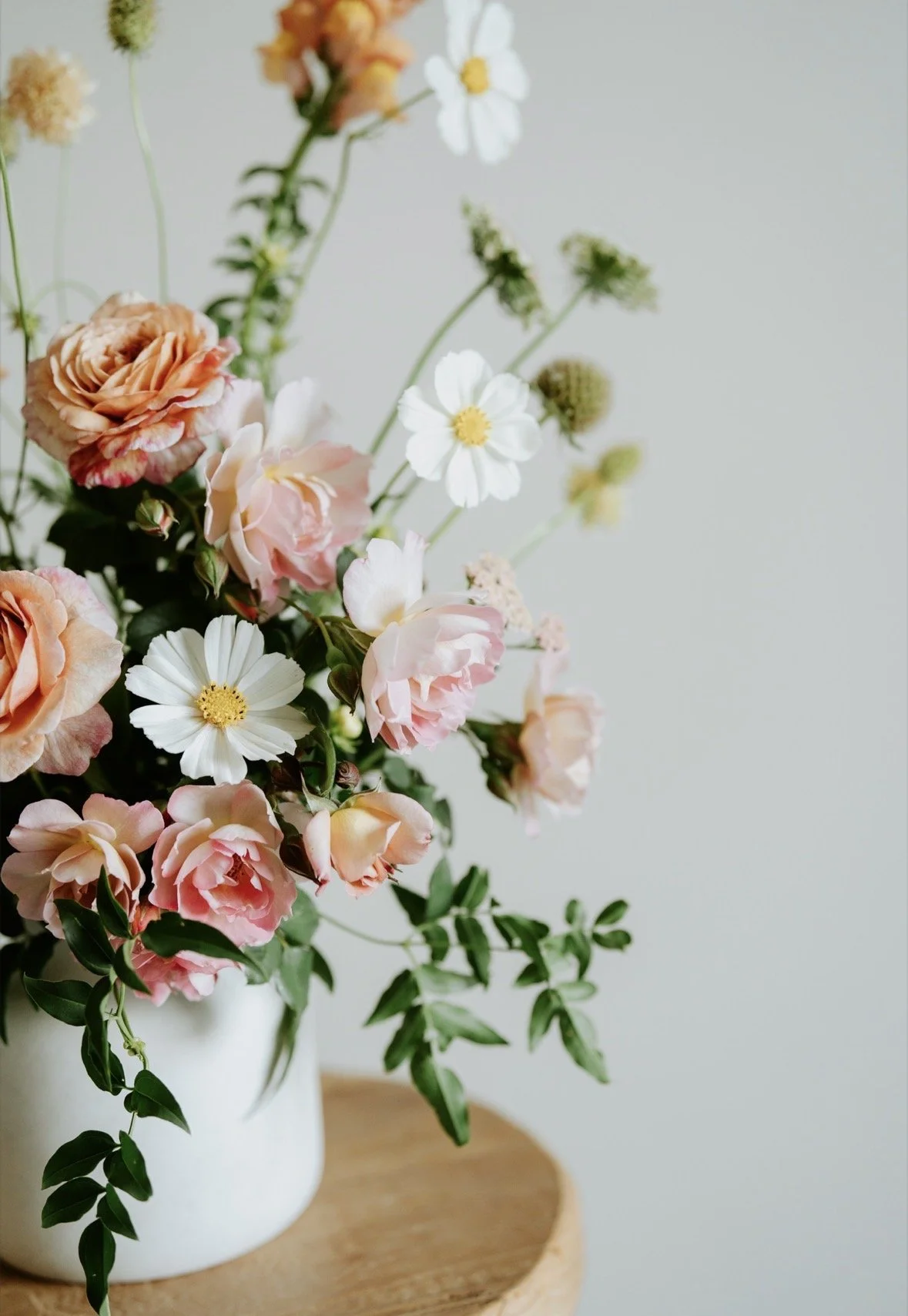 Close up of a bouquet of flowers in a white vase. Flowers are white and peach, with greenery.