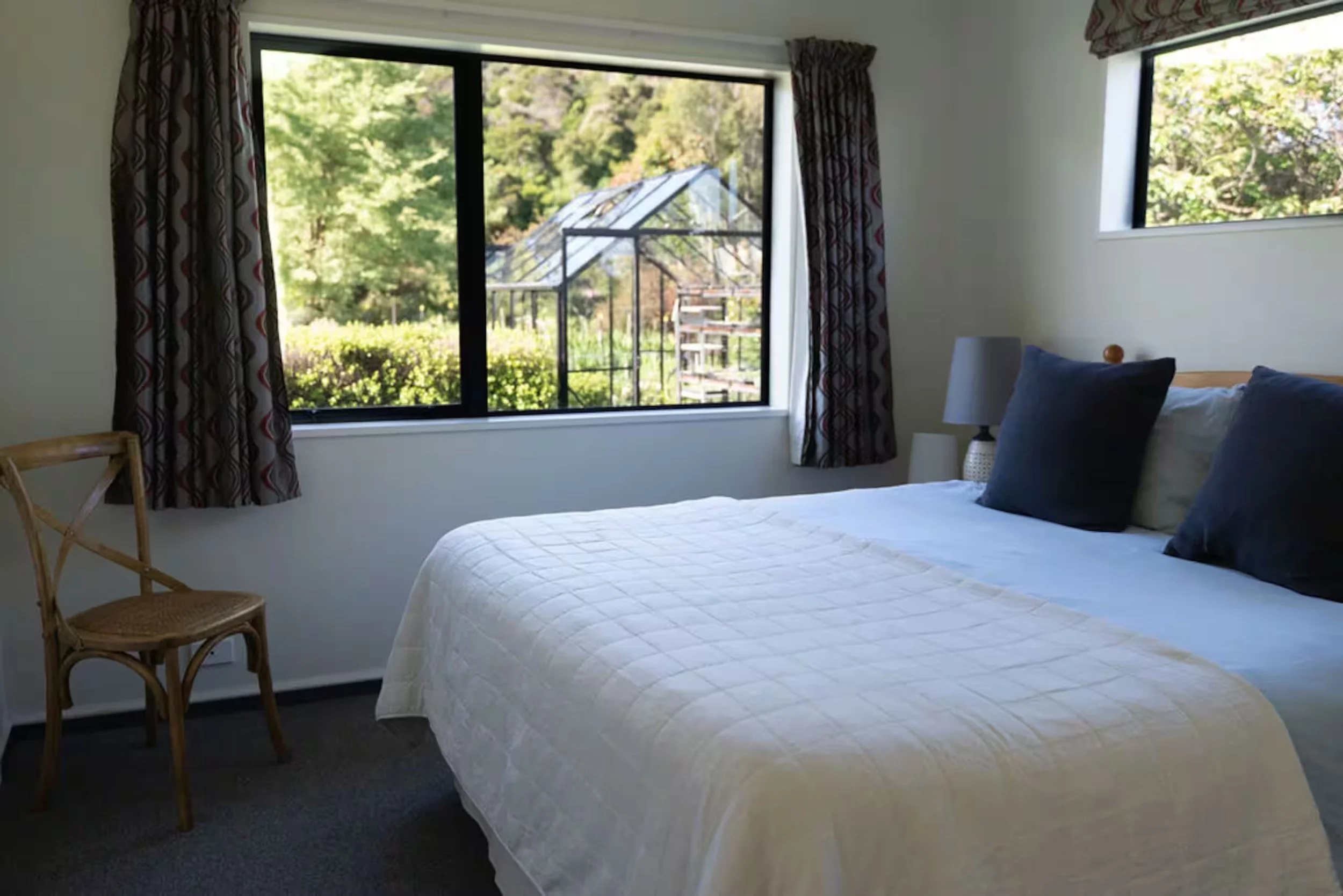 Bright bedroom with a white bed, navy pillows, patterned curtains, wicker chair, and garden greenhouse visible through the windows
