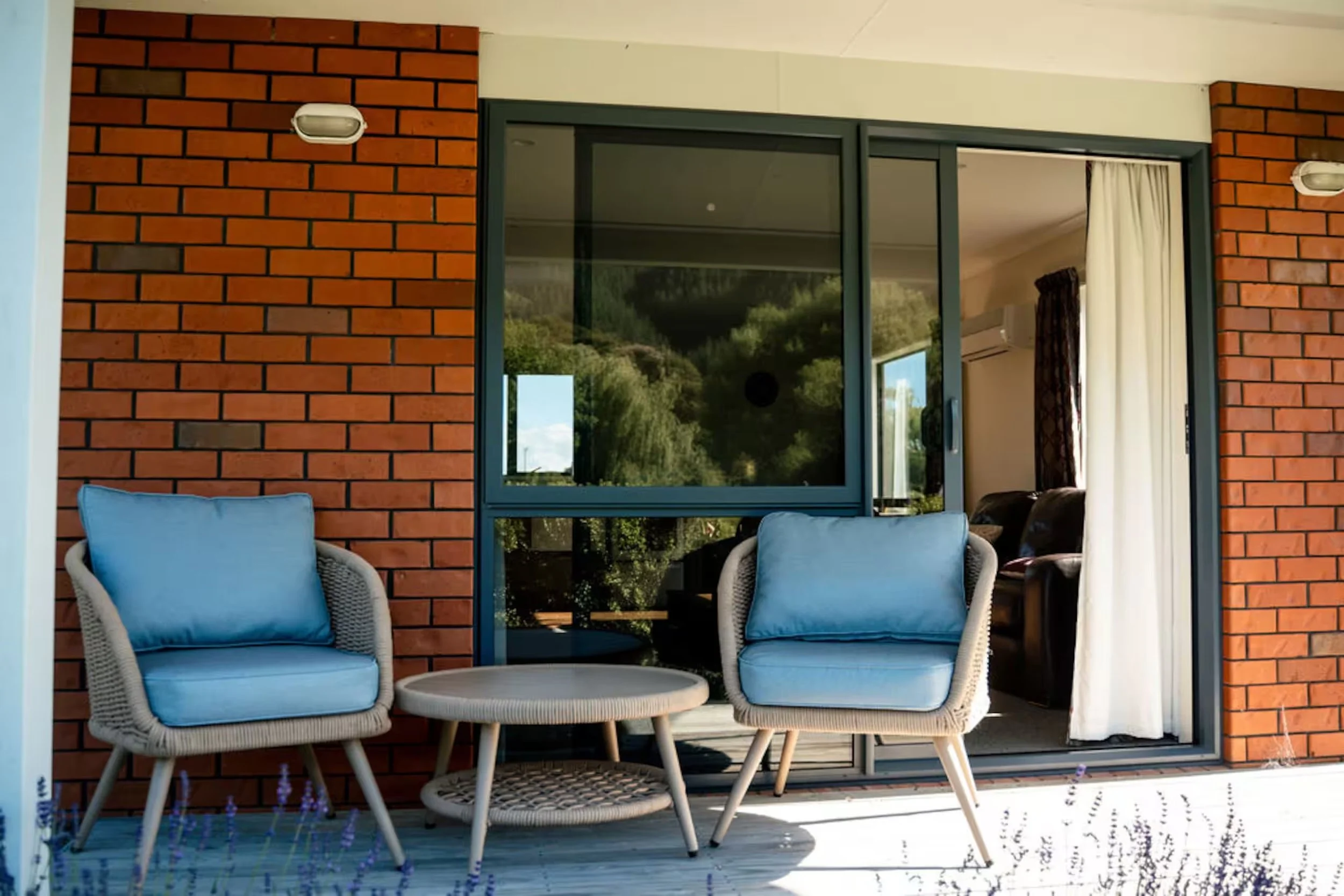 Outdoor patio with two blue-cushioned wicker chairs and a round table beside a brick house and glass doors