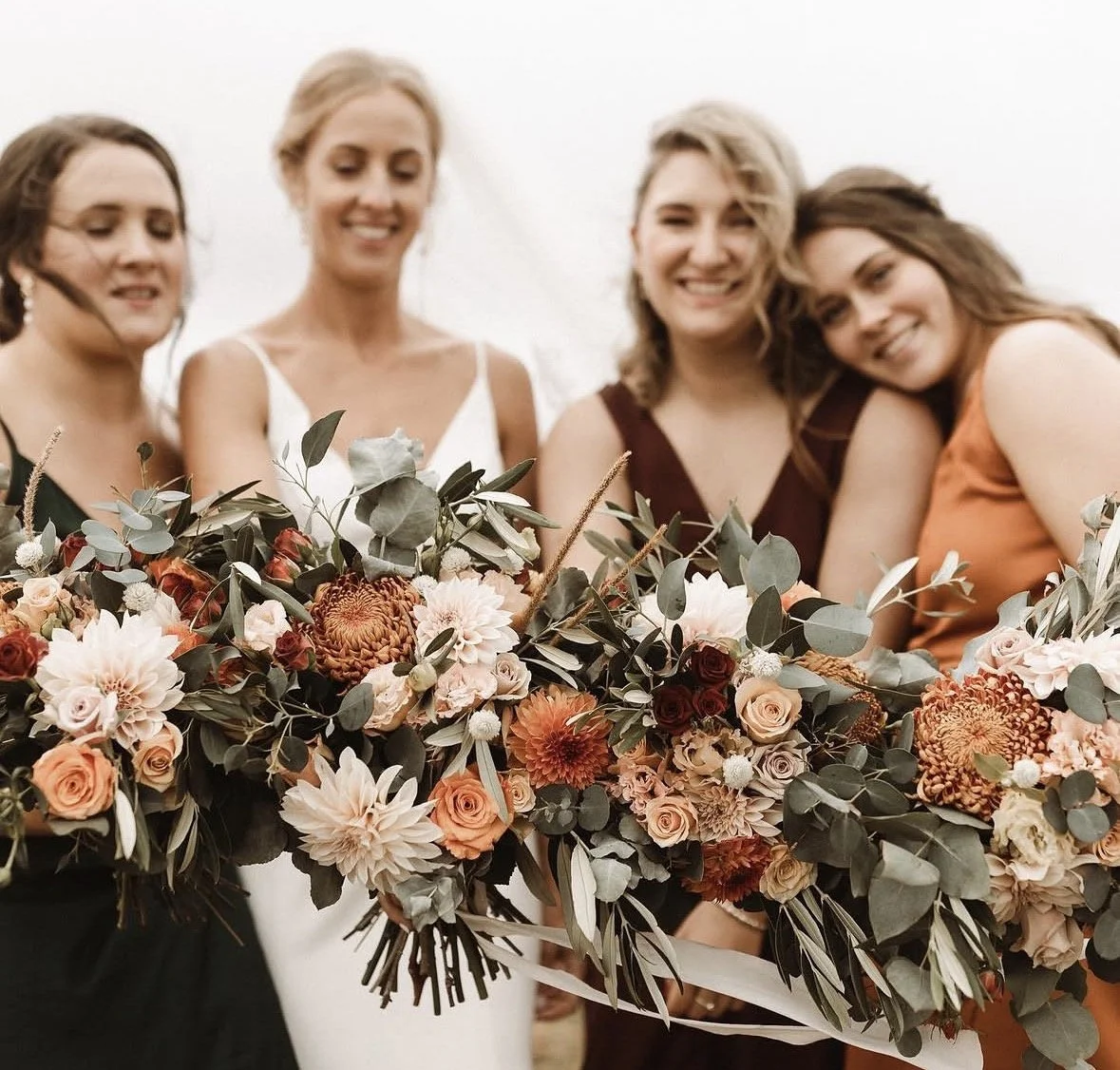 Bride and bridesmaids standing in a line showing off each of their bouquets. The bouquets include orange, peach, red, and white flowers with greenery to fill them out.