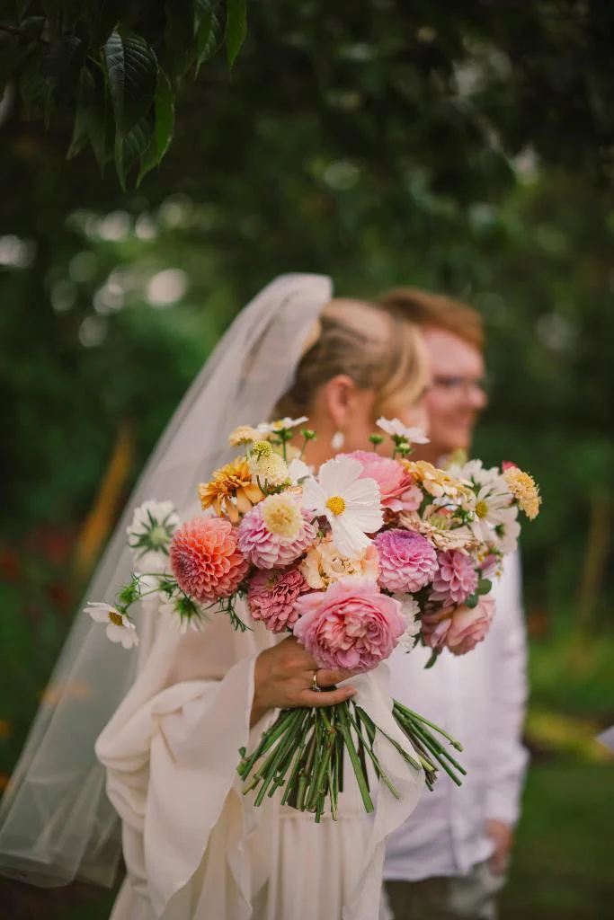 Bride holding a large pink and white flower bouquet outdoors, with her partner and veil softly blurred in the background