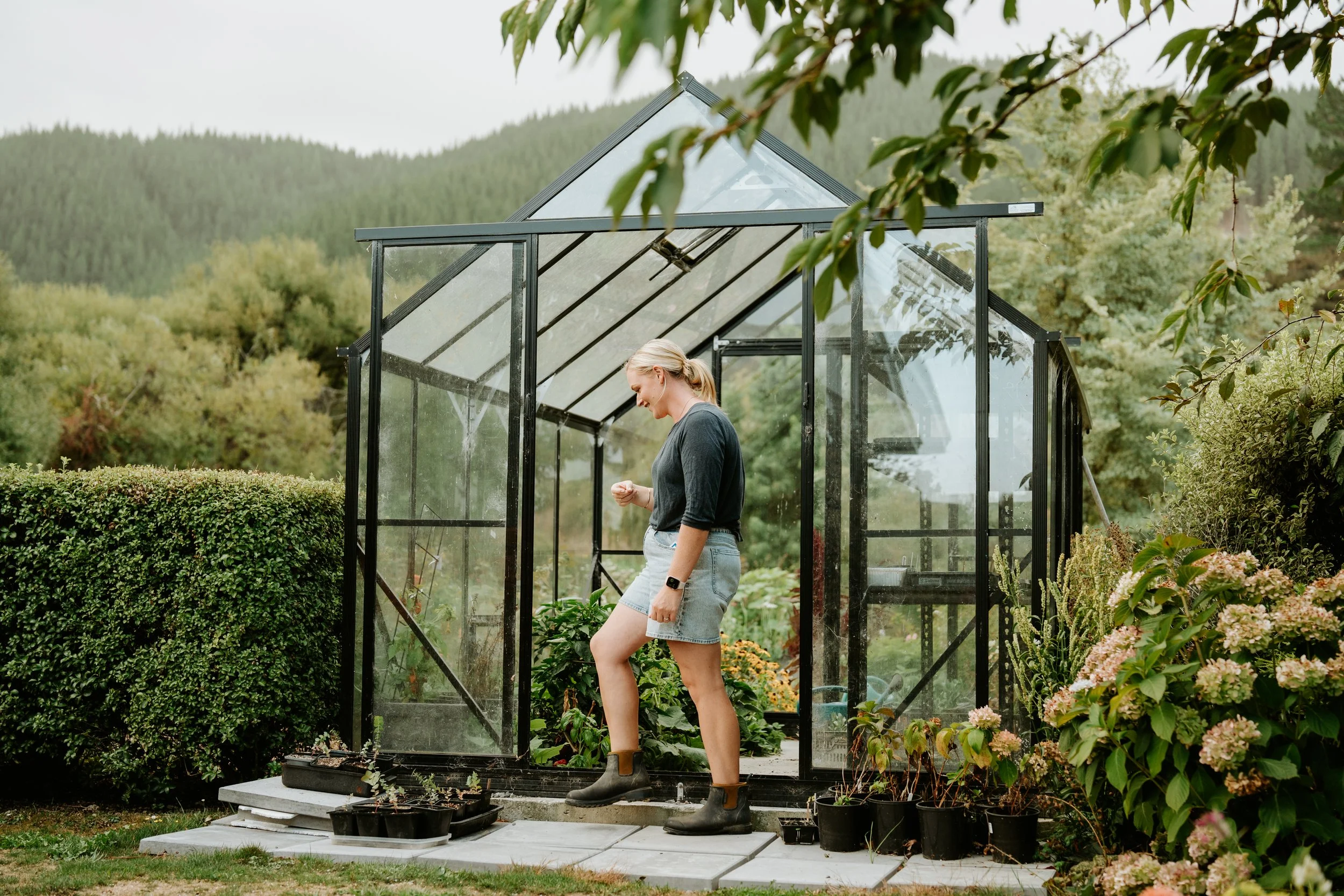 Woman in gardening boots standing inside a glass greenhouse surrounded by plants and forested hills