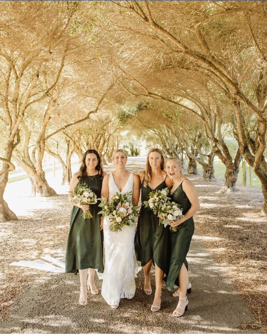 Bride and three bridesmaids holding bouquets under a tree-lined outdoor wedding aisle