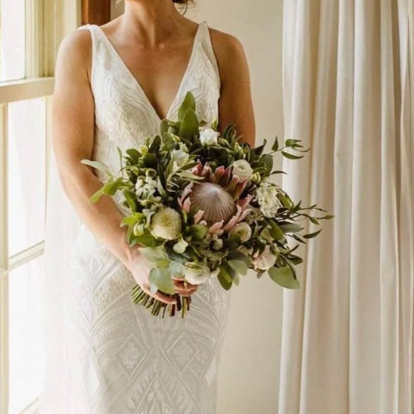 Bride in a white lace wedding dress holding a lush greenery and protea bouquet