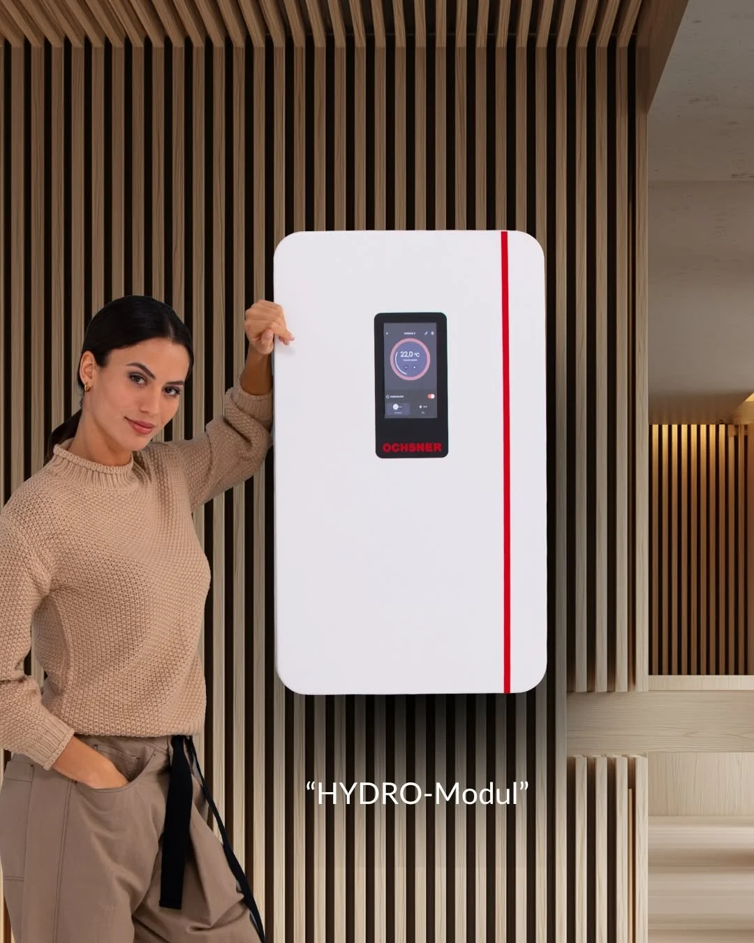 Eine Frau steht neben einer weißen Wandheizung mit Digitaldisplay, das die Temperatur anzeigt. Die Wandheizung trägt den Namen 'HYDRO-Modul'.