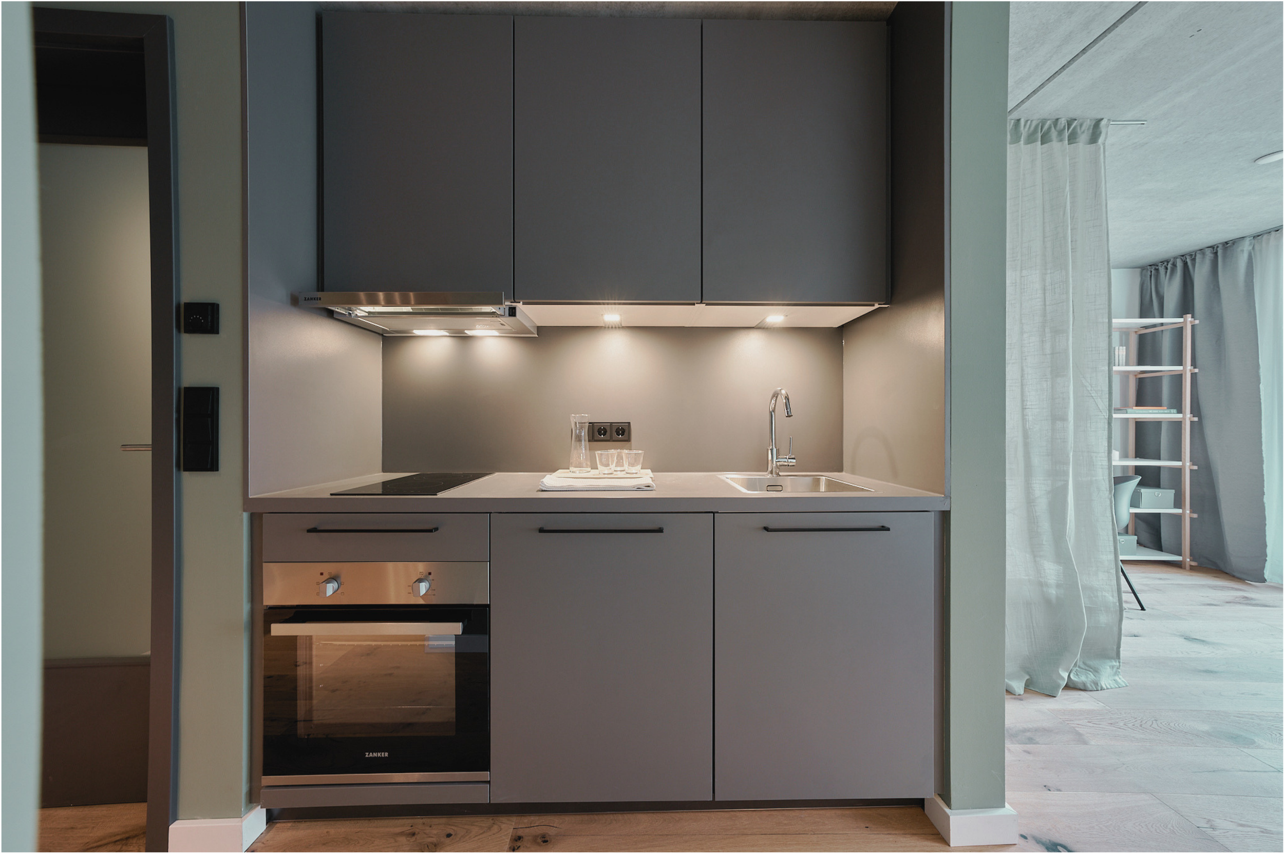 Moderne Küchenzeile mit grauen Schränken, eingebautem Backofen, Herd und Wasserhahn, im Hintergrund ein Wohnzimmer mit Vorhängen und Bücherregal.
