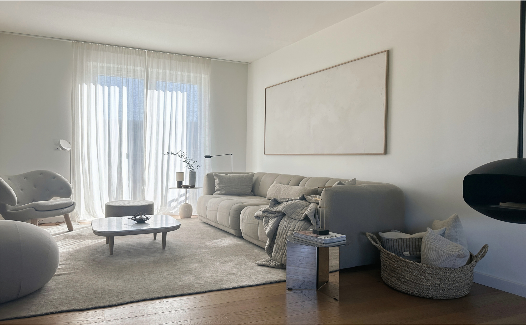 Modernes Wohnzimmer mit großen Fenstern, weißen Vorhängen, beige Couch, weißen Sesseln, Couchtisch, Korb mit Decke, kleiner Beistelltisch mit Büchern und Dekorationen, schwarze Stehlampe, abstraktes Kunstwerk an der Wand