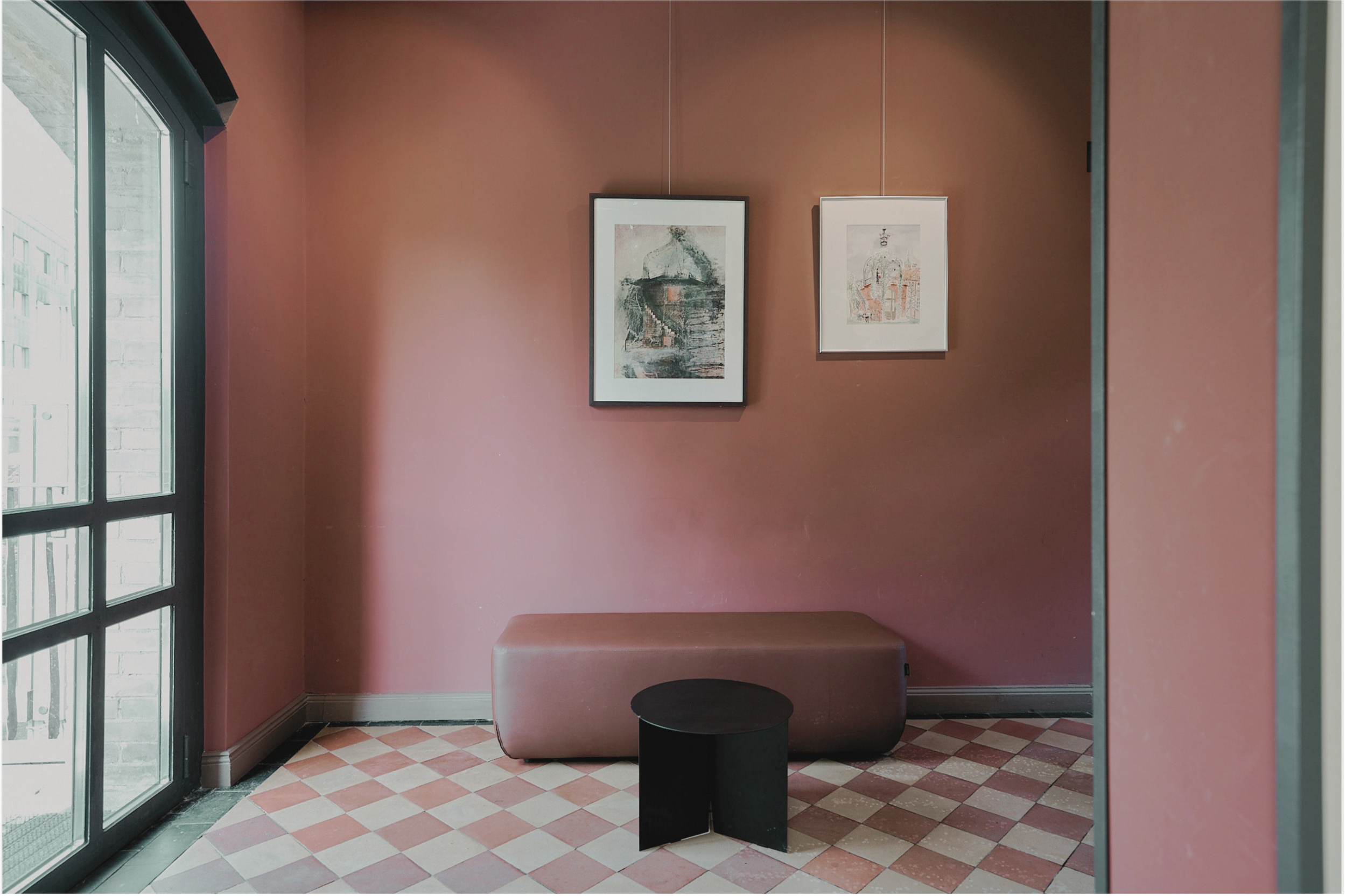 Ein Raum mit rosa Wänden, einem Fenster auf der linken Seite, auf dem Boden Fliesen in einem rot-weißen Muster. An der Wand hängen zwei Bilder, eines in einem schwarzen Rahmen, das andere in einem weißen Rahmen. Ein gepolsteter Sitz oder Bank ist unter den Bildern, mit einem kleinen schwarzen Tisch davor.