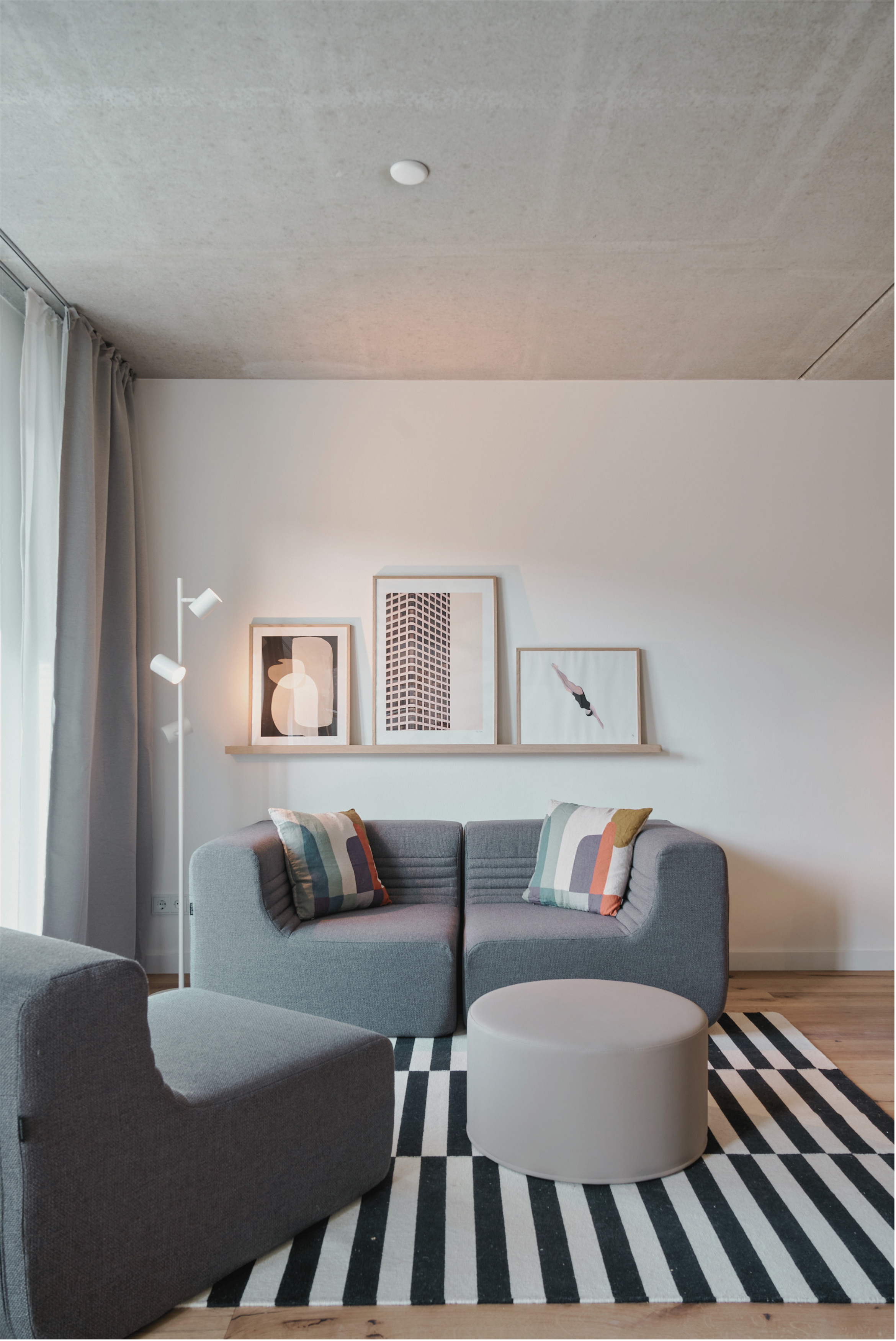 Wohnzimmer mit grauer Couch, bunten Kissen, schwarzen und weißen Streifen Teppich, weißer runder Couchtisch, Wandkunst mit abstrakten Bildern, Stehlampe, Gardinen, Decke mit einem Rauchmelder.