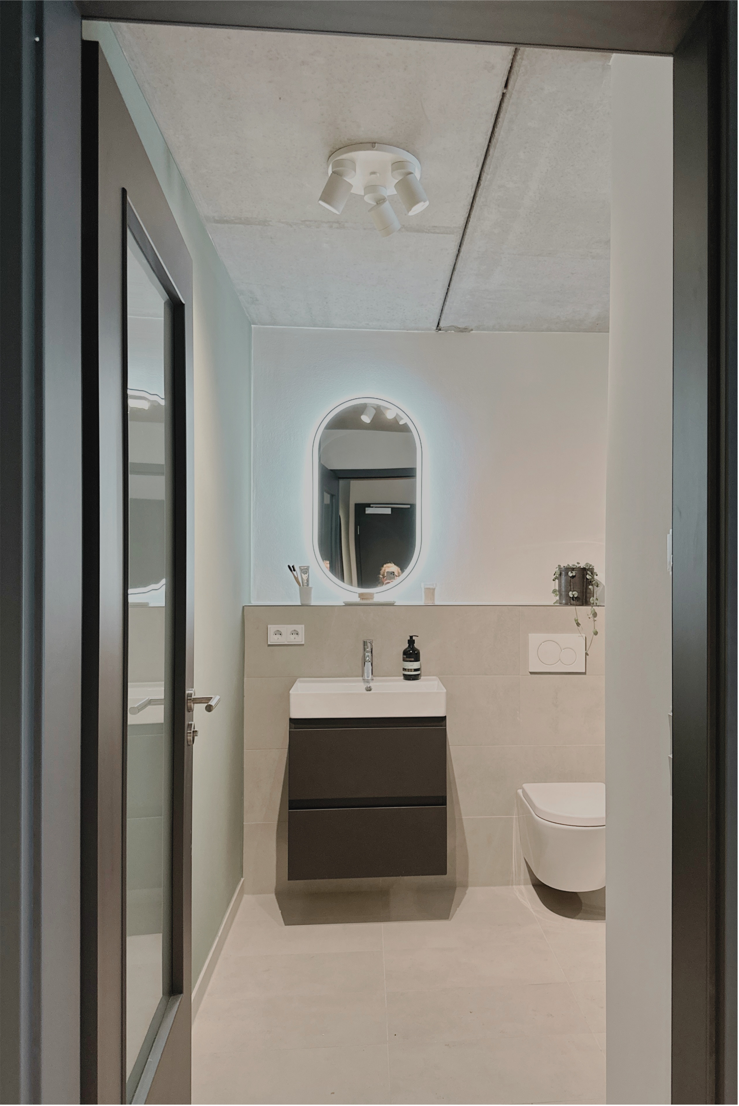 Ein moderner Badezimmerraum mit Spiegel, Waschbecken, Badmöbeln und Toilette, helles Design mit neutralen Farben.