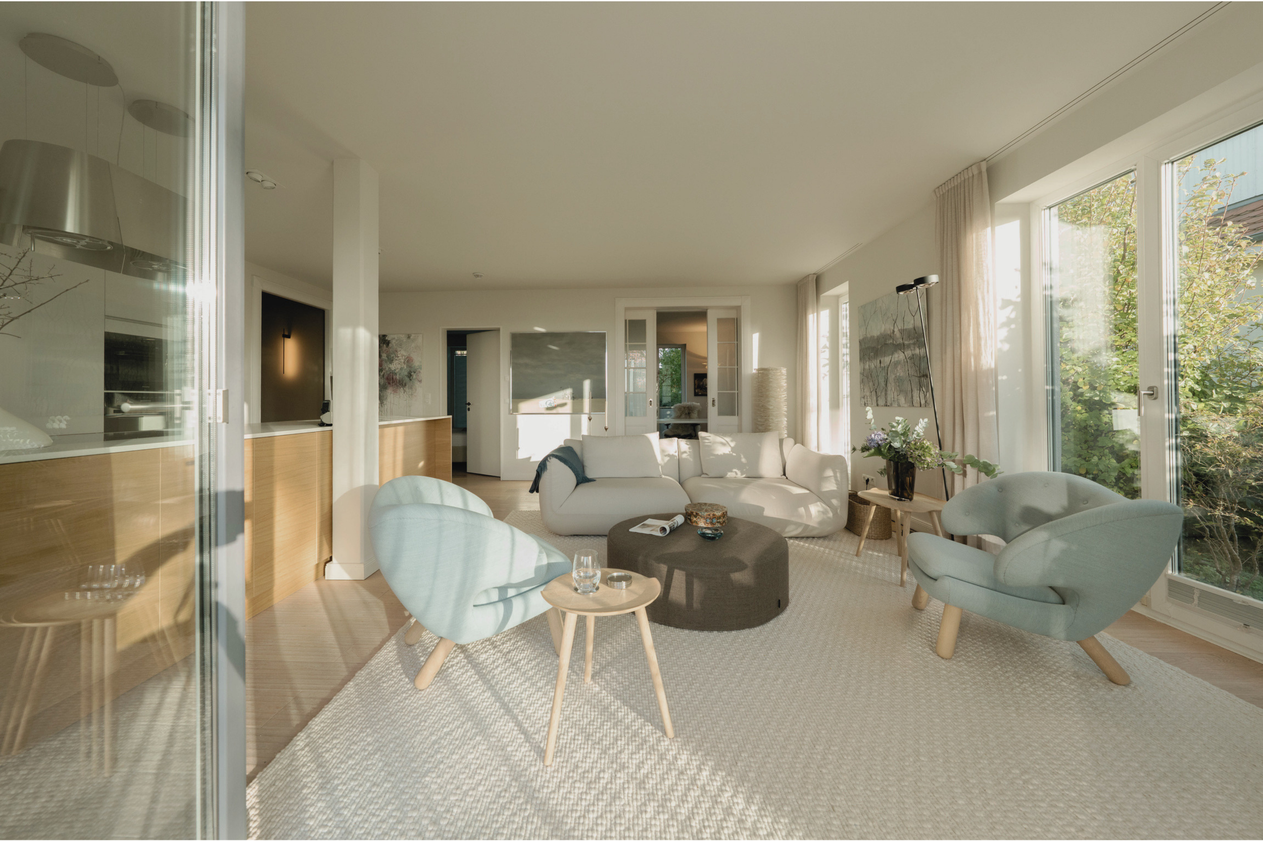 Ein hell eingerichteter Wohnzimmerraum mit großen Fenstern, einer beigen Couch, zwei hellblauen Sesseln, einem runden grauen Couchtisch, einer Lampe und Wandkunst, bei Tageslicht.