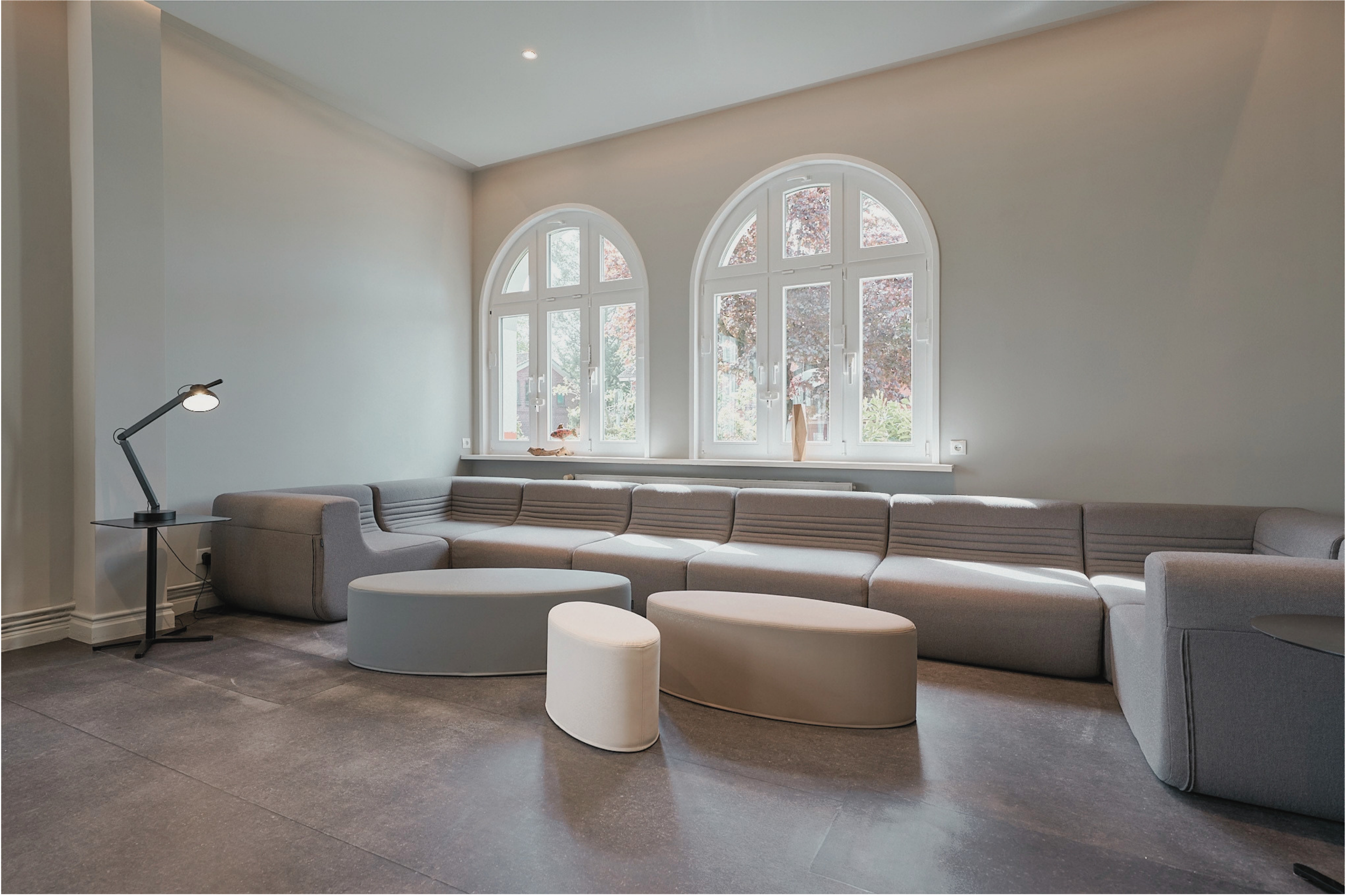 Modernes Wohnzimmer mit großen, weißen, halbkreisförmigen Fenstern, grauem Sofa und passenden Sitzgelegenheiten, Stehlampe, dunklem Boden und weißer Wand