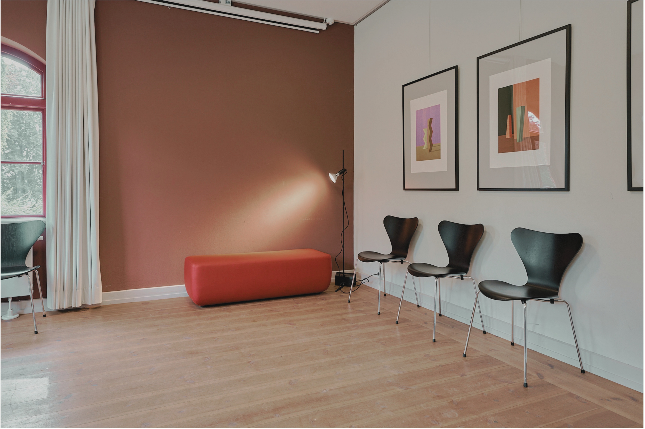 Wartezimmer mit mehreren schwarzen Stühlen, einem roten Hocker, einem Stehlampe und Bildern an der Wand.